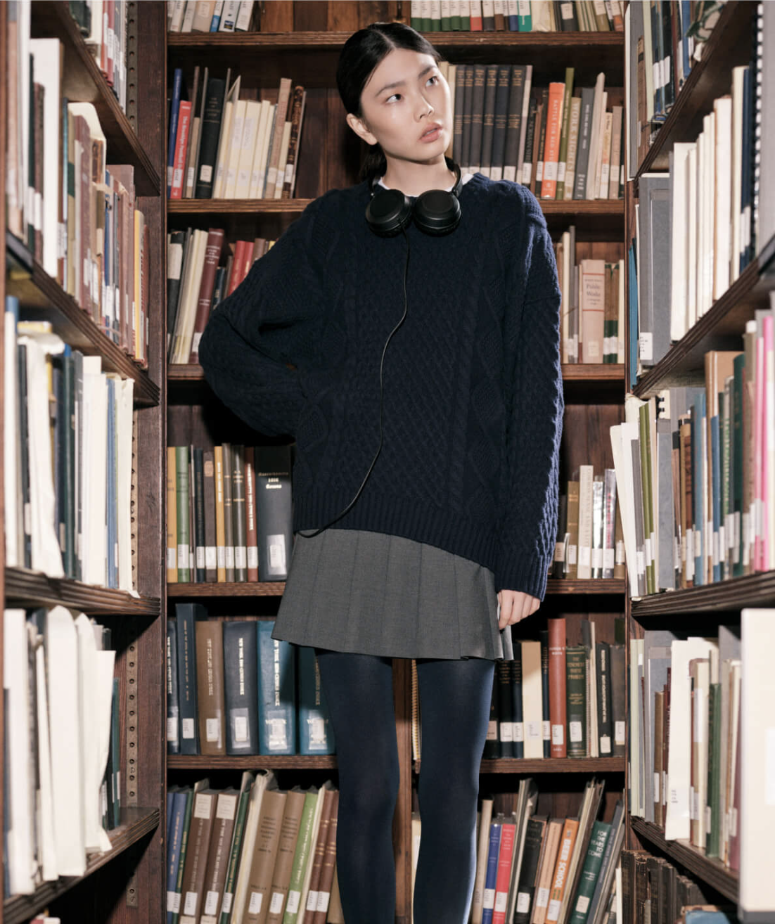 A young woman stands between bookshelves in a library, wearing a navy blue sweater, gray pleated skirt, black tights, and headphones around her neck, looking contemplatively to her left.