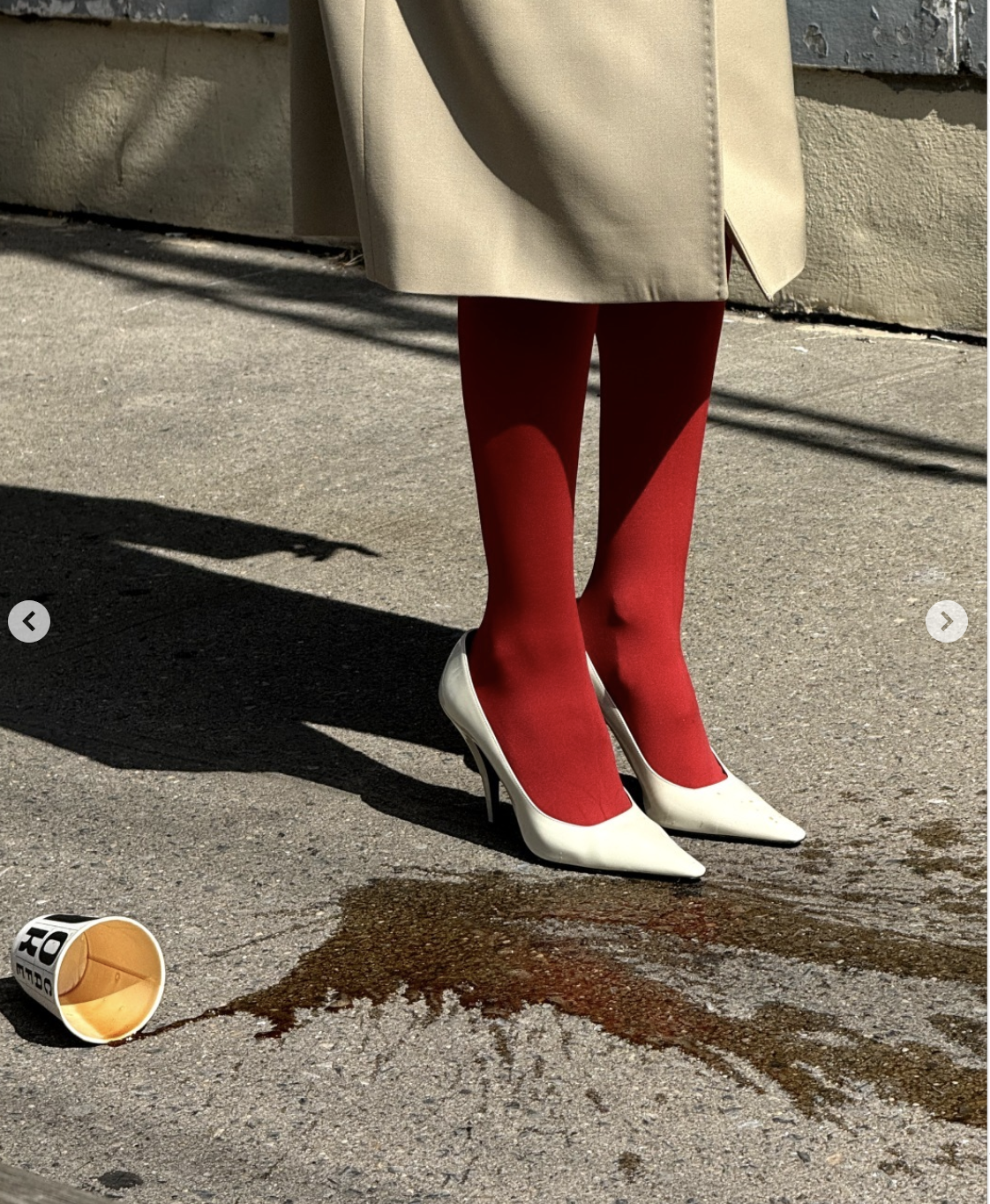 A person wearing red stockings, white high heels, a beige coat, and the bottom portion of their outfit standing outdoors on pavement with spilled coffee nearby.