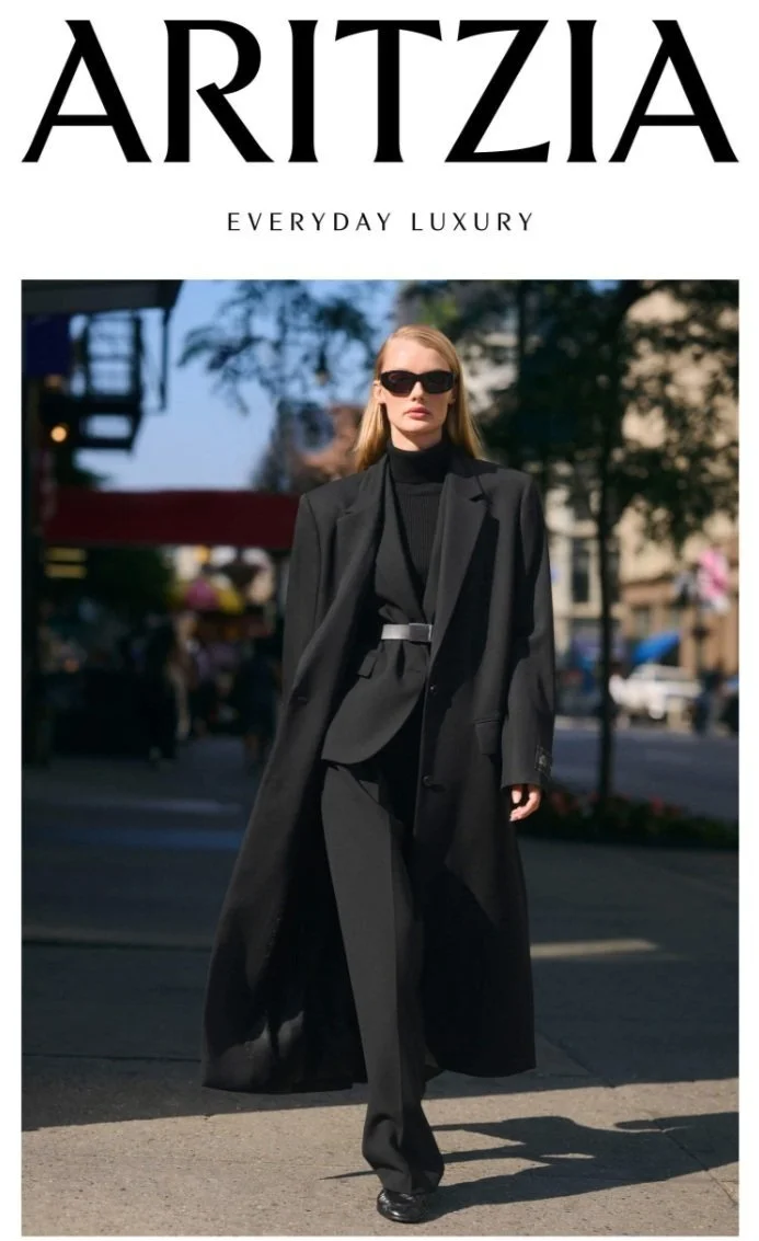 A woman walking on a city street wearing a black suit with a long coat, black pants, and sunglasses, with trees and buildings in the background.