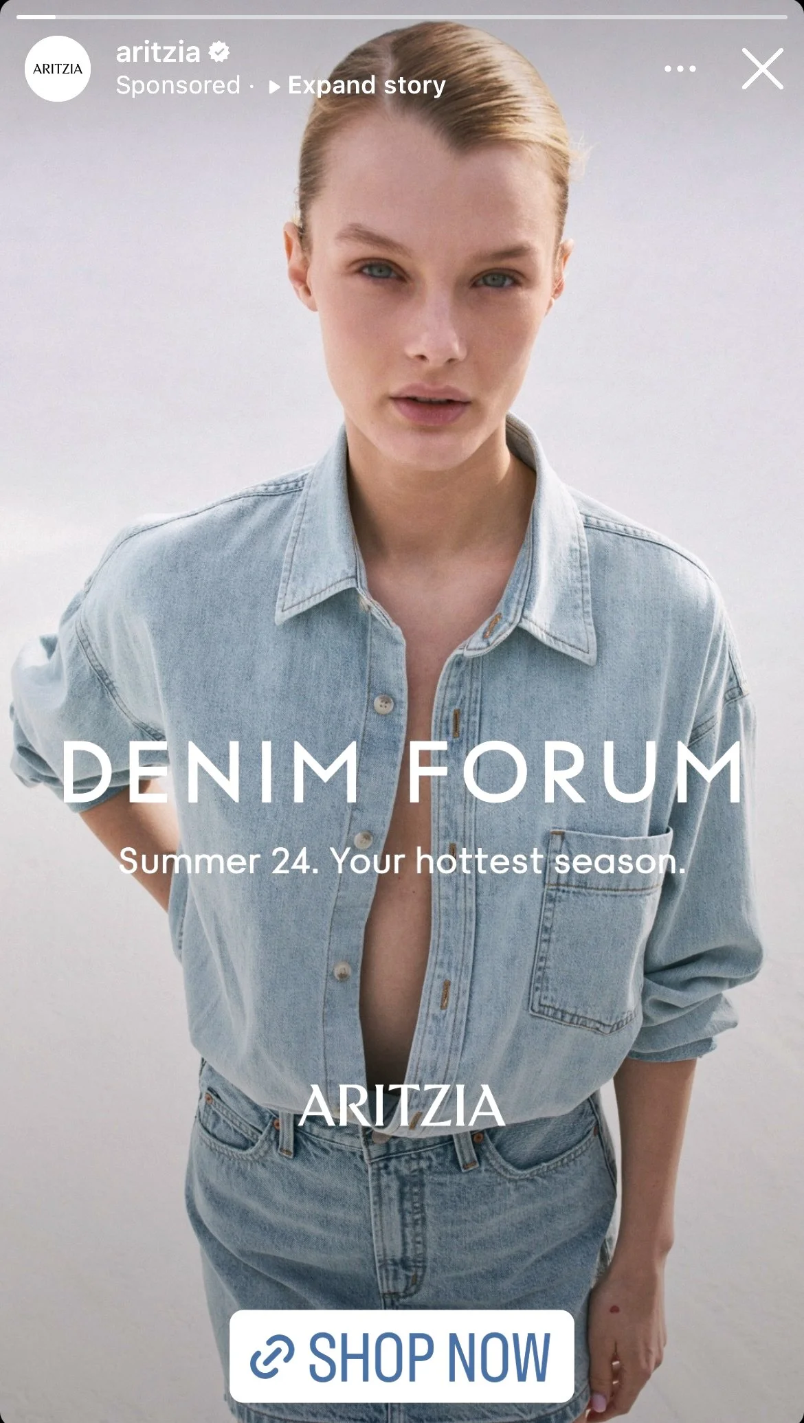 A woman with short blonde hair wearing a light blue denim shirt and jeans, standing against a light background.