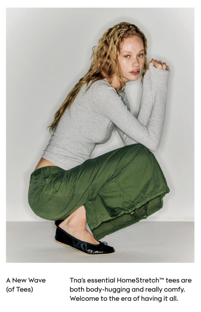 A woman with long, wavy blonde hair is squatting against a plain white background. She is wearing a light gray long-sleeve top, loose-fitting green cargo pants, and black flats. She has a relaxed expression and is looking slightly to the side.