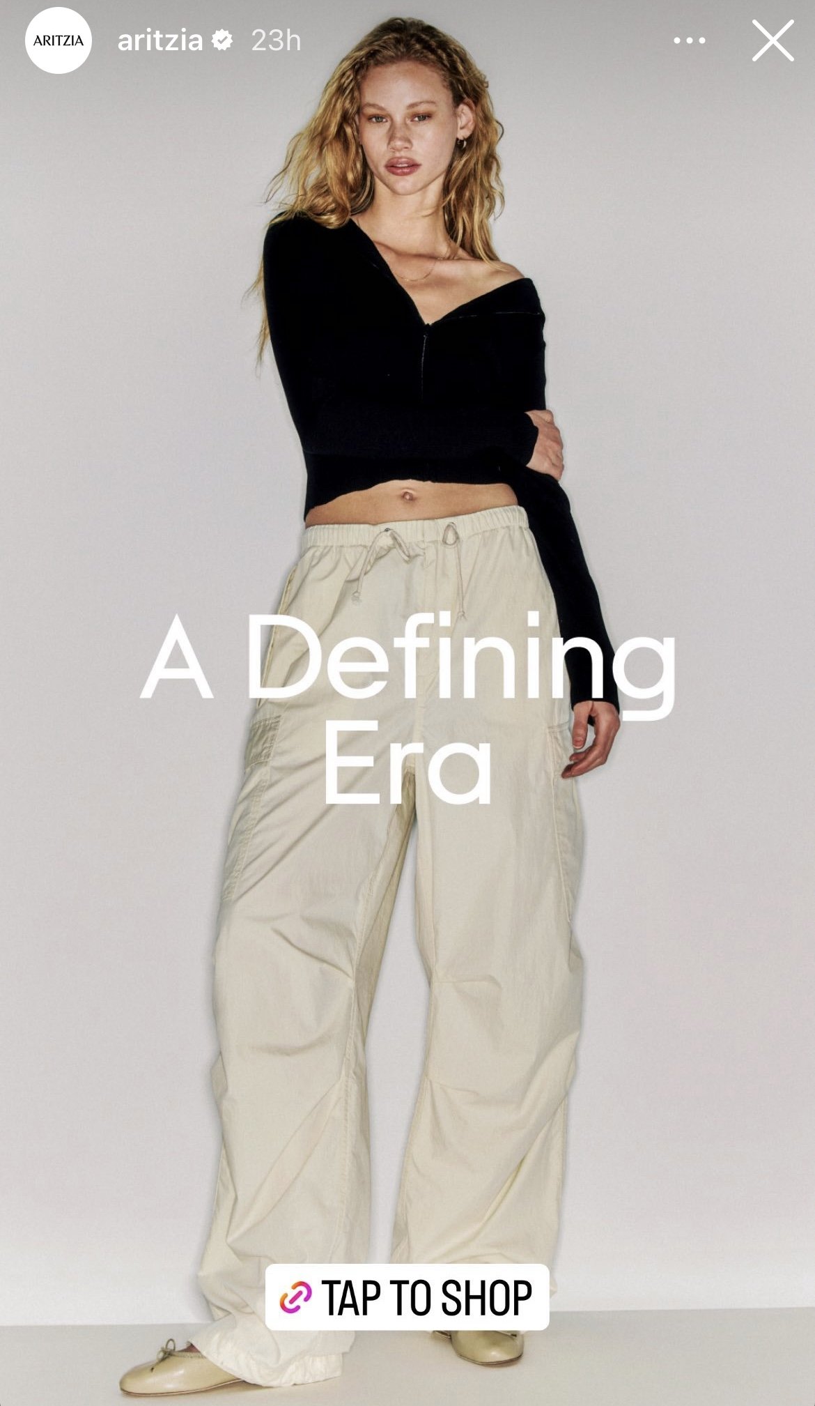 A young woman with wavy red hair wearing a black off-shoulder top and beige wide-leg pants, standing against a plain background with the text "A Defining Era" and a "TAP TO SHOP" button overlay.