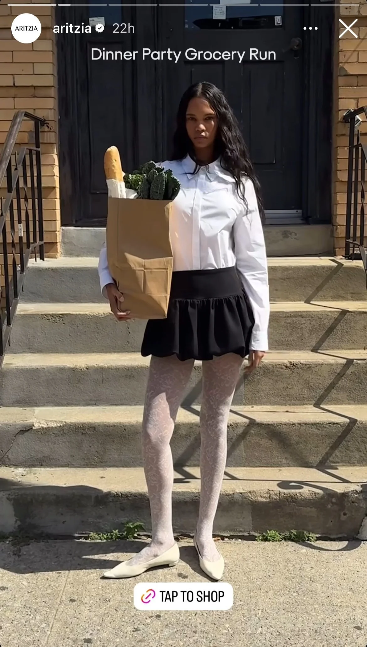 A young woman standing outside on stairs holding a grocery bag with produce, wearing a white shirt, black skirt, white lace tights, and white shoes. The background shows a brick building and a black door.