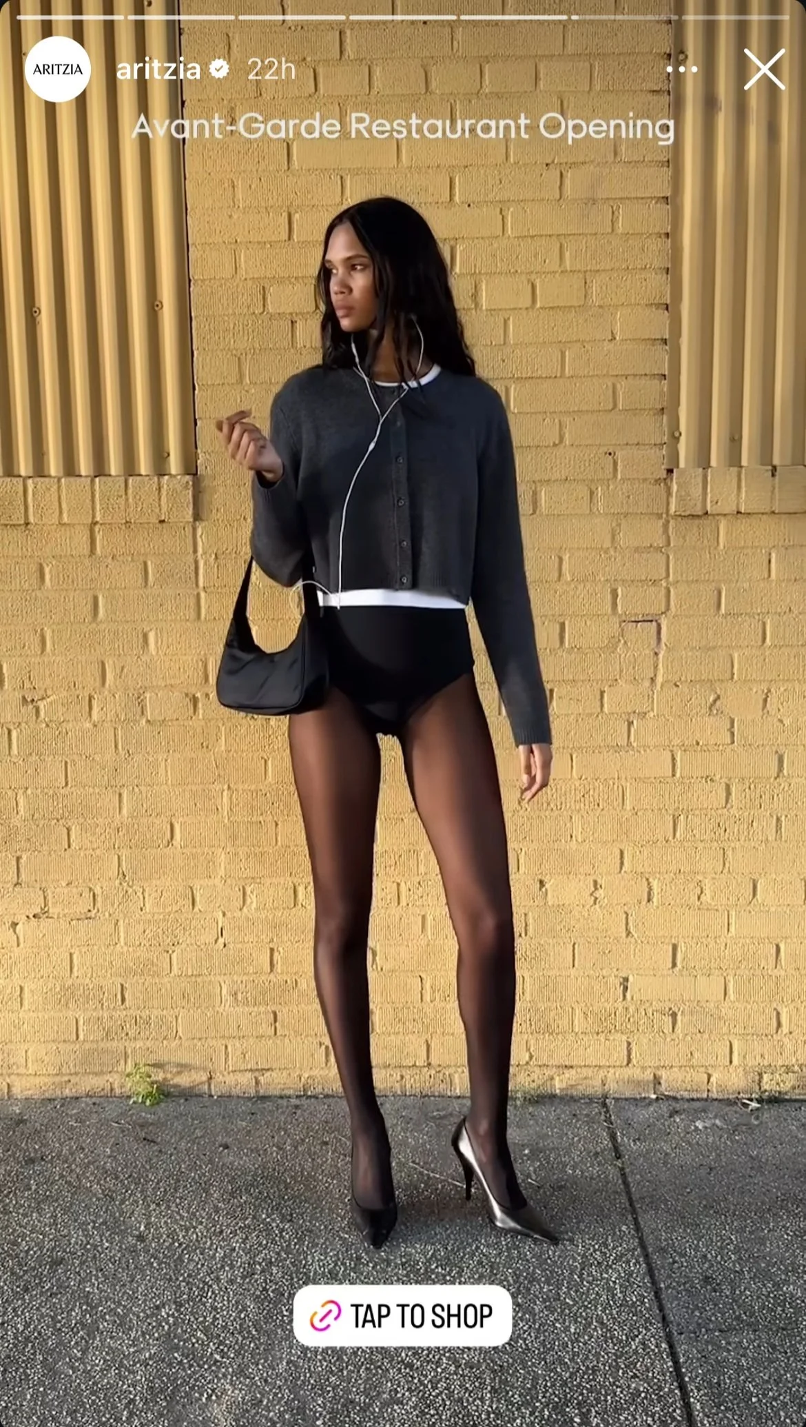 A woman standing against a yellow brick wall, wearing sheer black pantyhose and black high heels, with a cropped gray sweater, black purse, and white shirt underneath, holding earphones.
