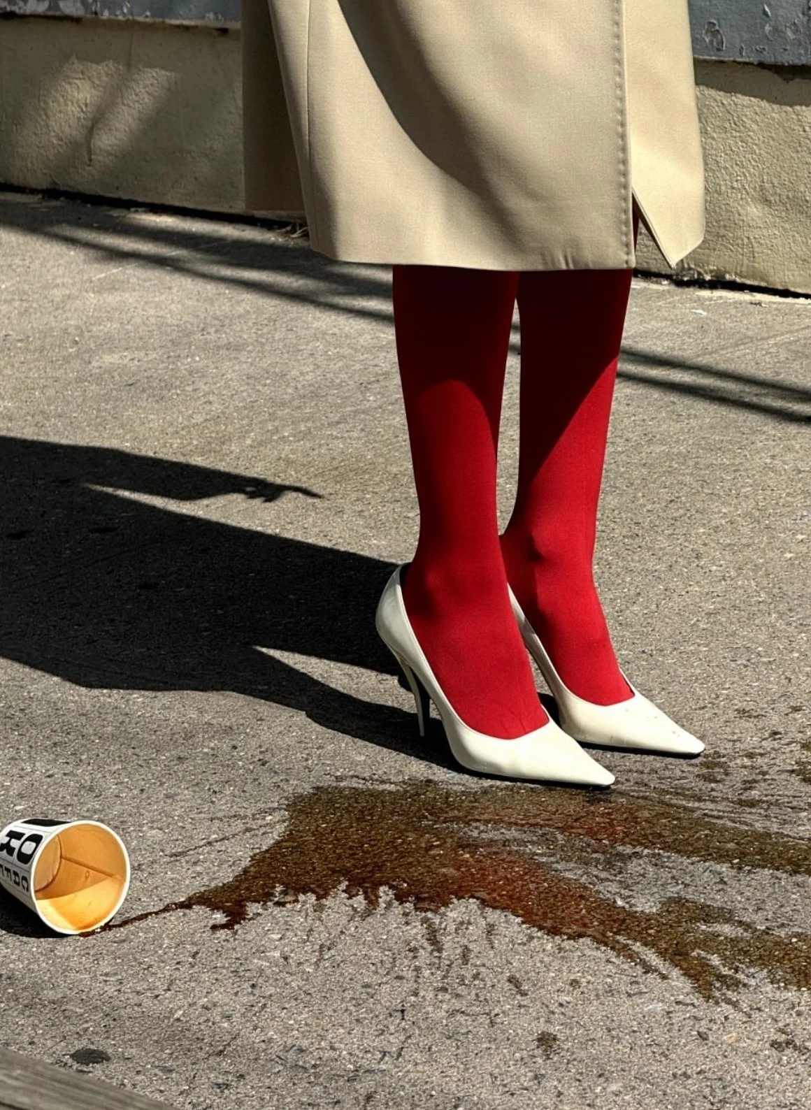 Close-up of a person standing on the street, wearing a beige coat, red tights, and white high-heeled shoes. There is spilled coffee or another dark liquid on the pavement near the person's feet, with some of the liquid spilling from an overturned paper cup.