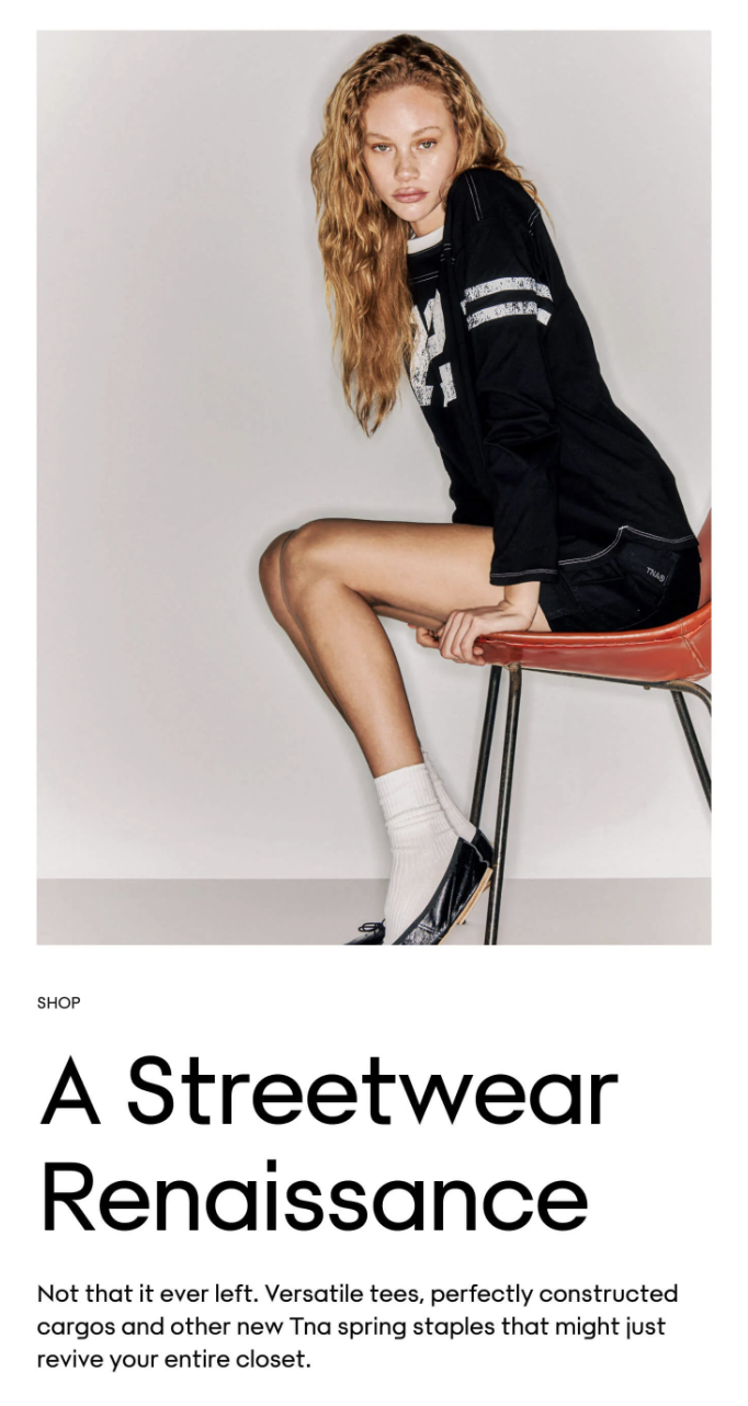 Young woman with curly blonde hair sitting on a red chair, wearing a black varsity jacket, white socks, and black shoes, against a plain white background.