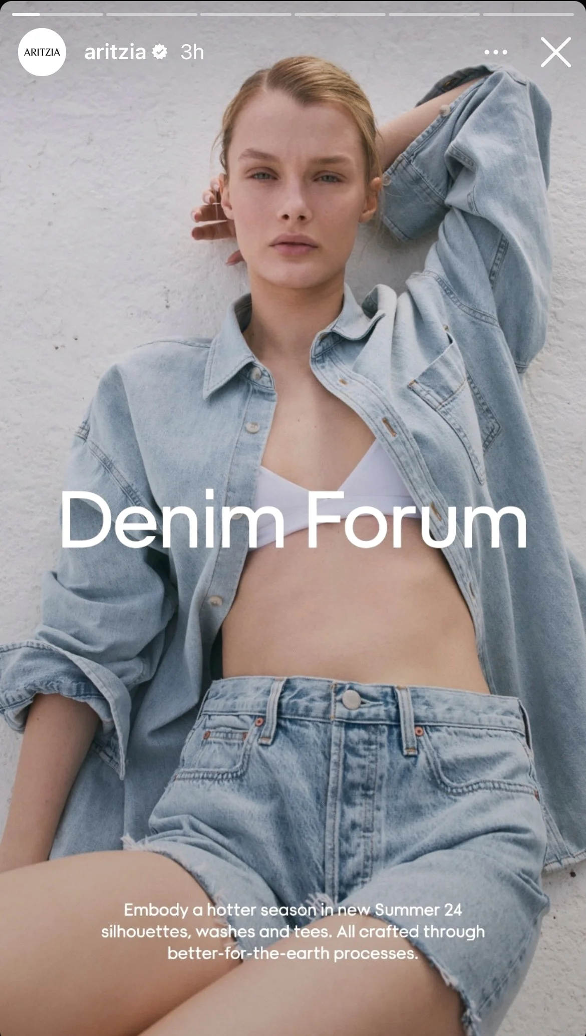 Young woman with fair skin, blonde hair, wearing a light denim jacket, white bralette, and high-waisted denim shorts, leaning against a white wall.
