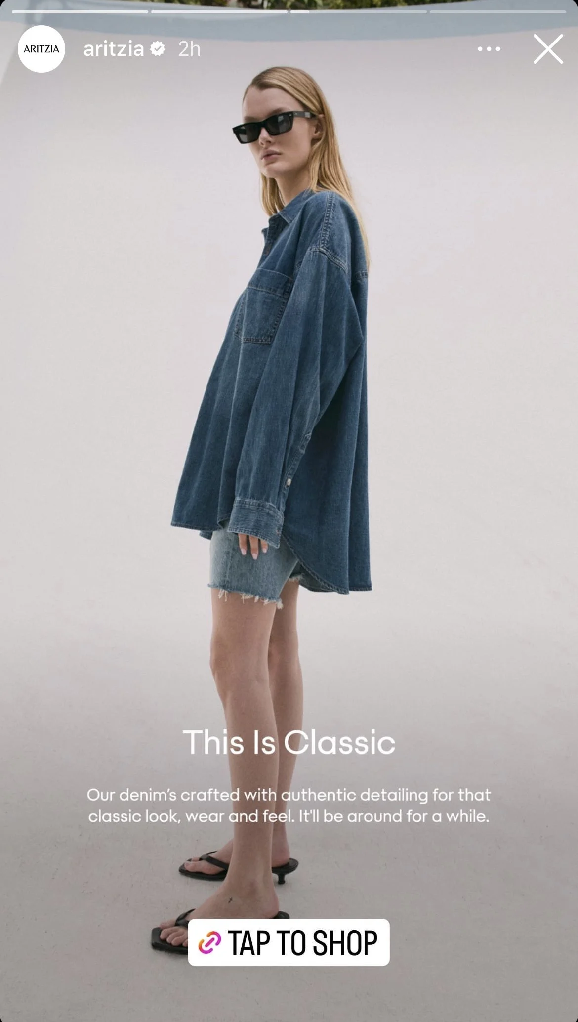 Woman with blonde hair wearing black sunglasses, oversized denim jacket, frayed denim shorts, black flip-flops, standing against a plain light background. Text overlay promoting classic denim with a tap-to-shop button.