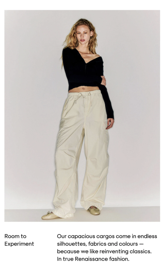 Young woman with curly blonde hair wearing a black cropped sweater and loose cream-colored cargo pants standing against a plain white background.