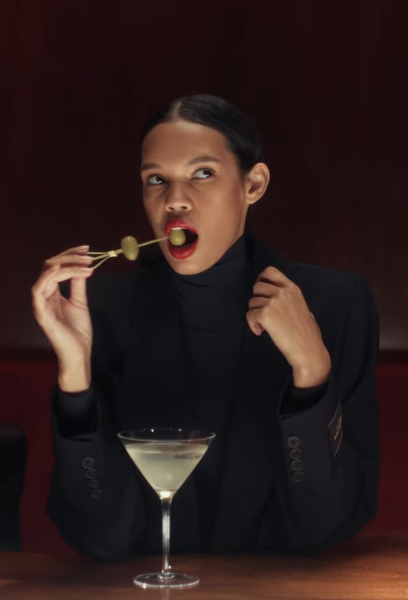 A woman with dark lipstick and slicked-back hair holding green olives on skewers, sitting at a table with a martini glass in front of her, making a playful expression with her eyes looking upward.
