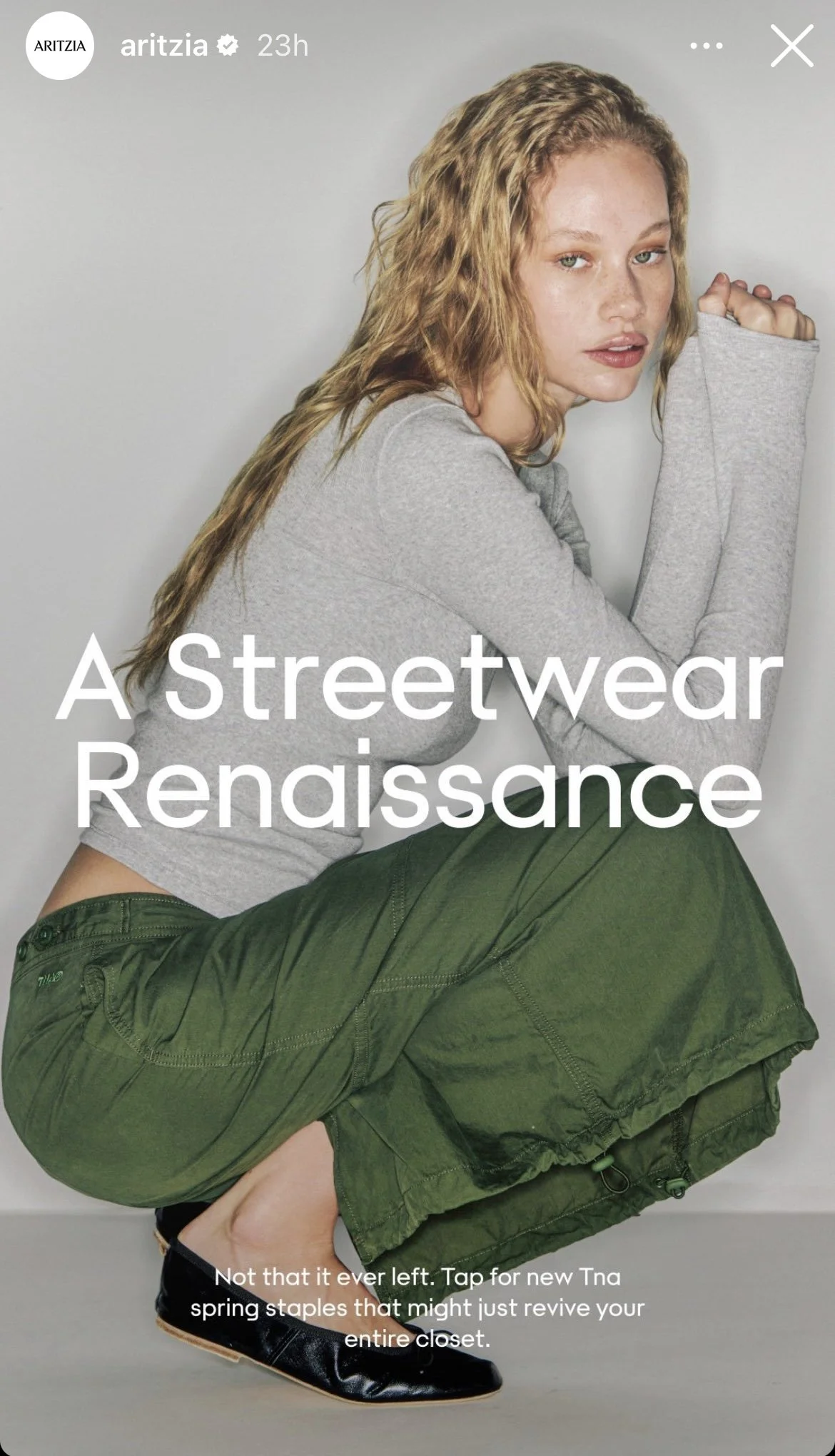 A woman with wavy red hair wearing a gray long-sleeve top and green streetwear pants, crouching against a plain background.