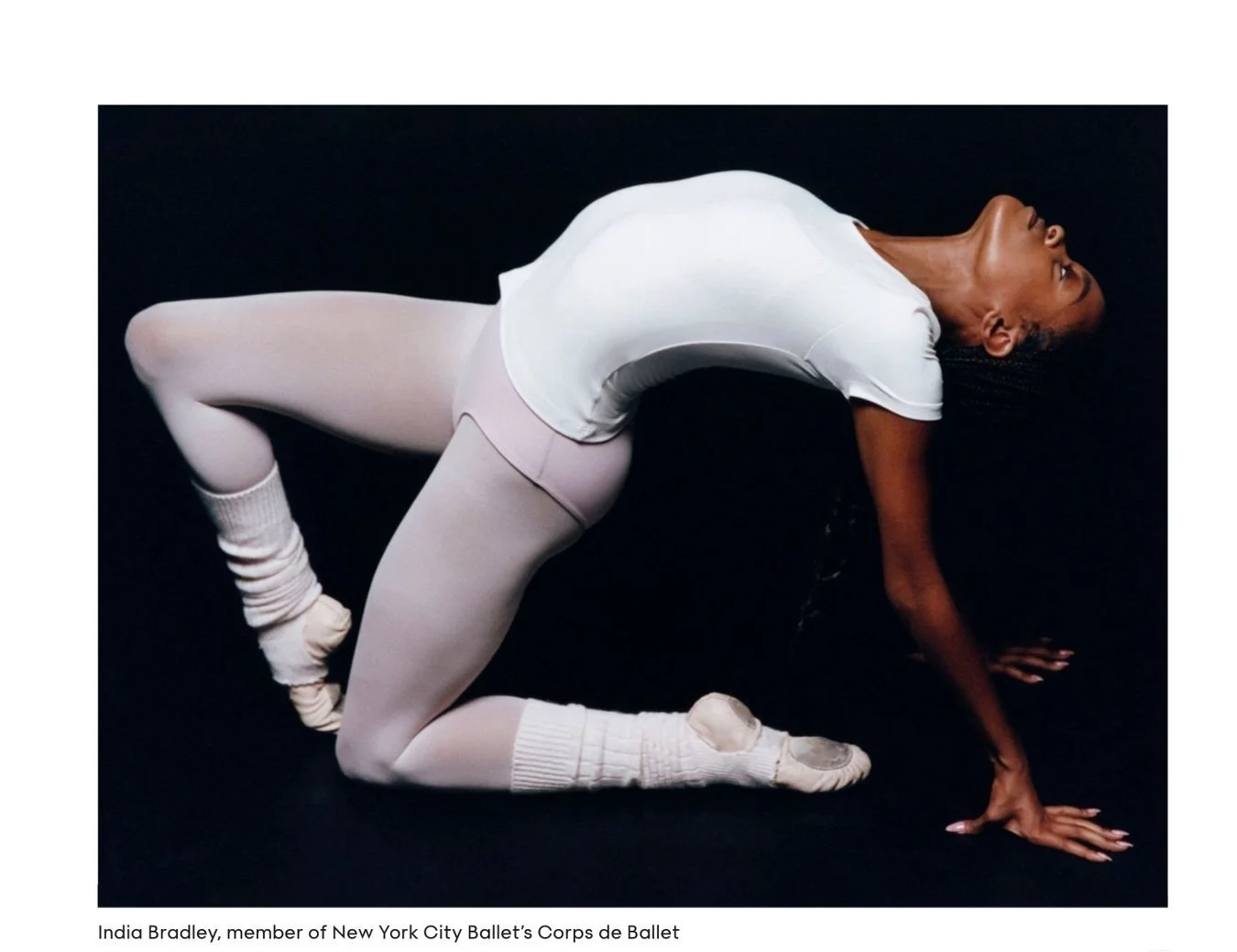 A dancer is on all fours on a black floor, wearing a white t-shirt, pink tights, and ballet slippers.