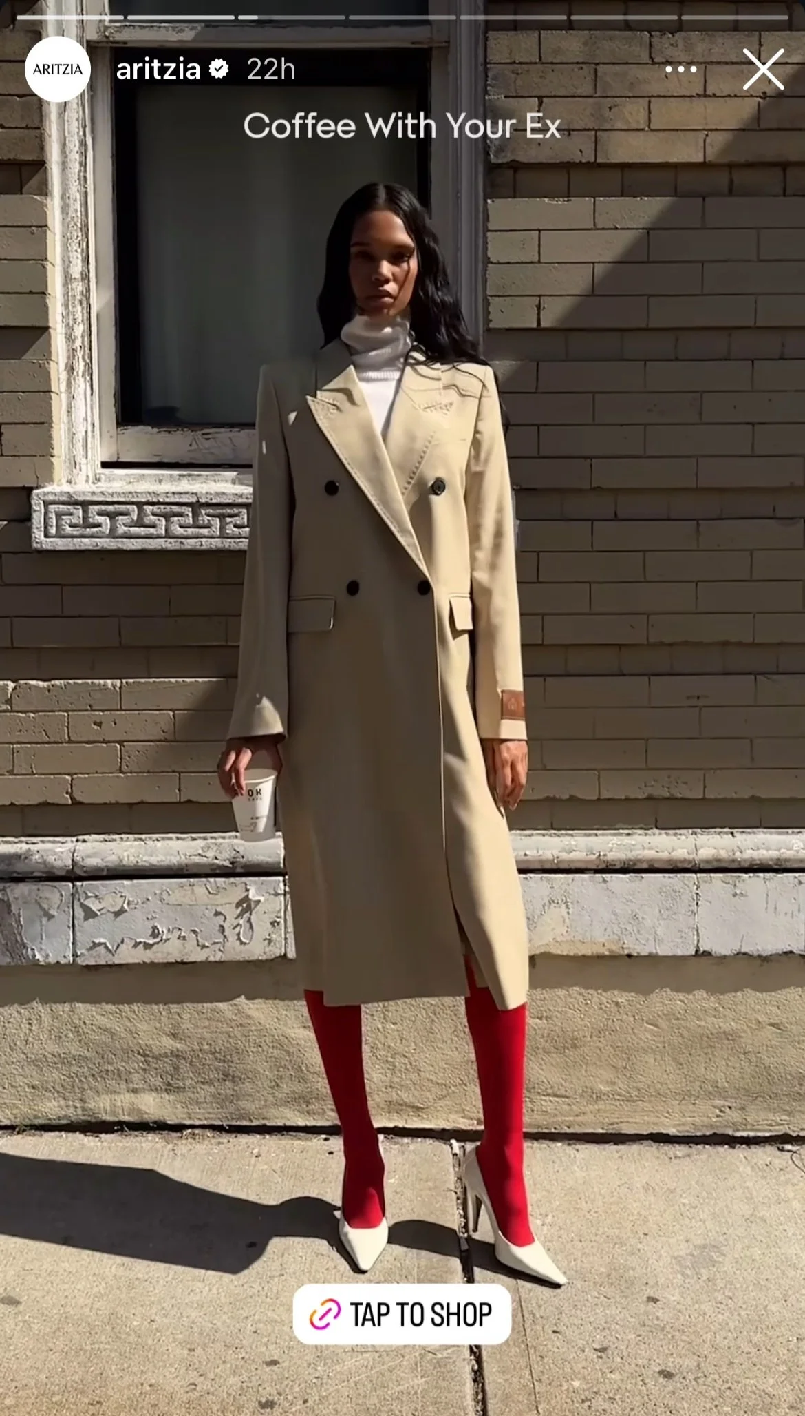 A woman standing outside in front of a brick wall, wearing a beige double-breasted trench coat, red stockings, and white high heels, holding a coffee cup.