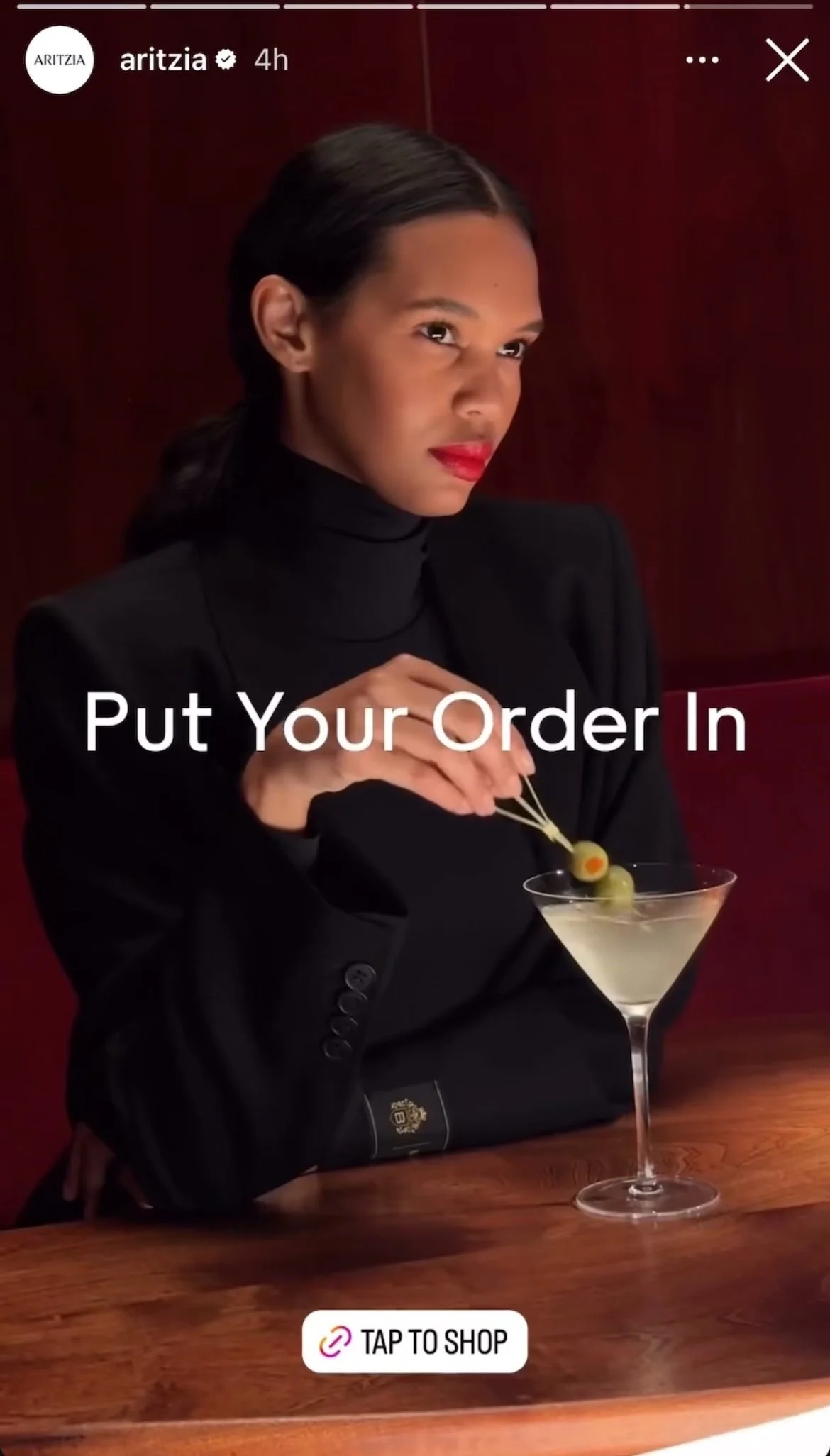 A woman dressed in black with red lipstick sitting at a wooden table, garnishing a martini with green olives on picks, with text overlay saying "Put Your Order In" and a "Tap to Shop" button.