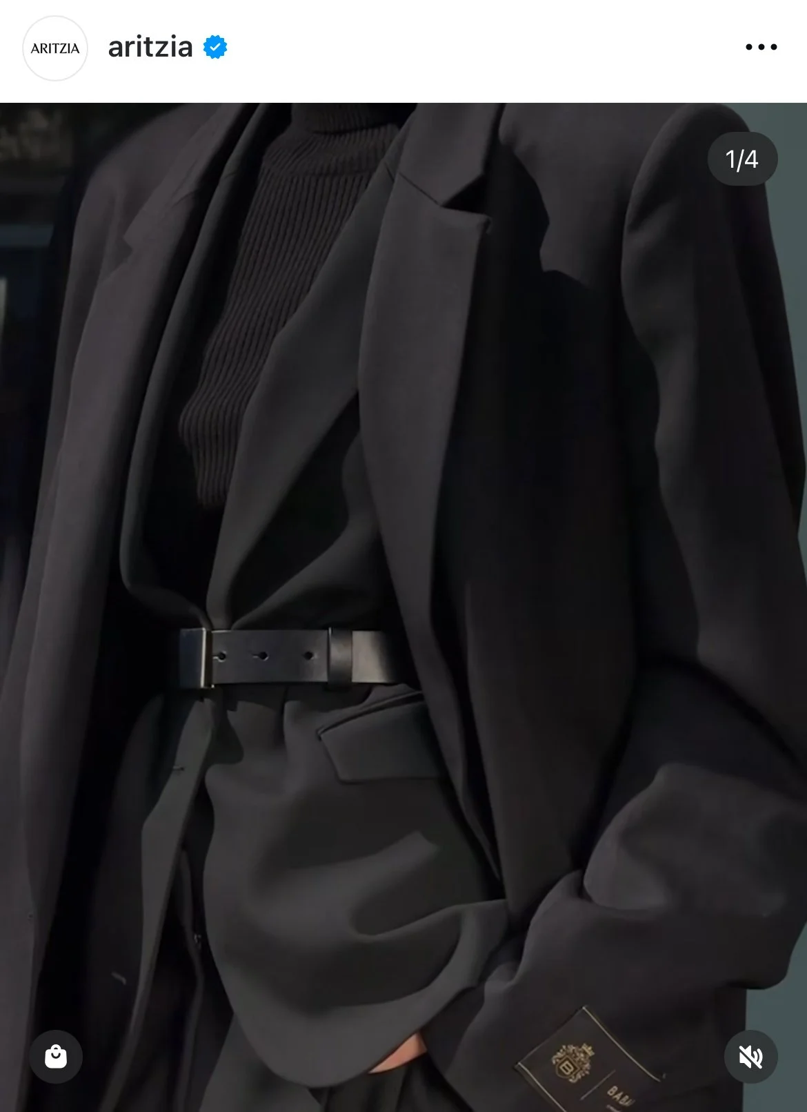 Close-up of a person dressed in black wearing a blazer, turtleneck, and belt, with a label on the sleeve.