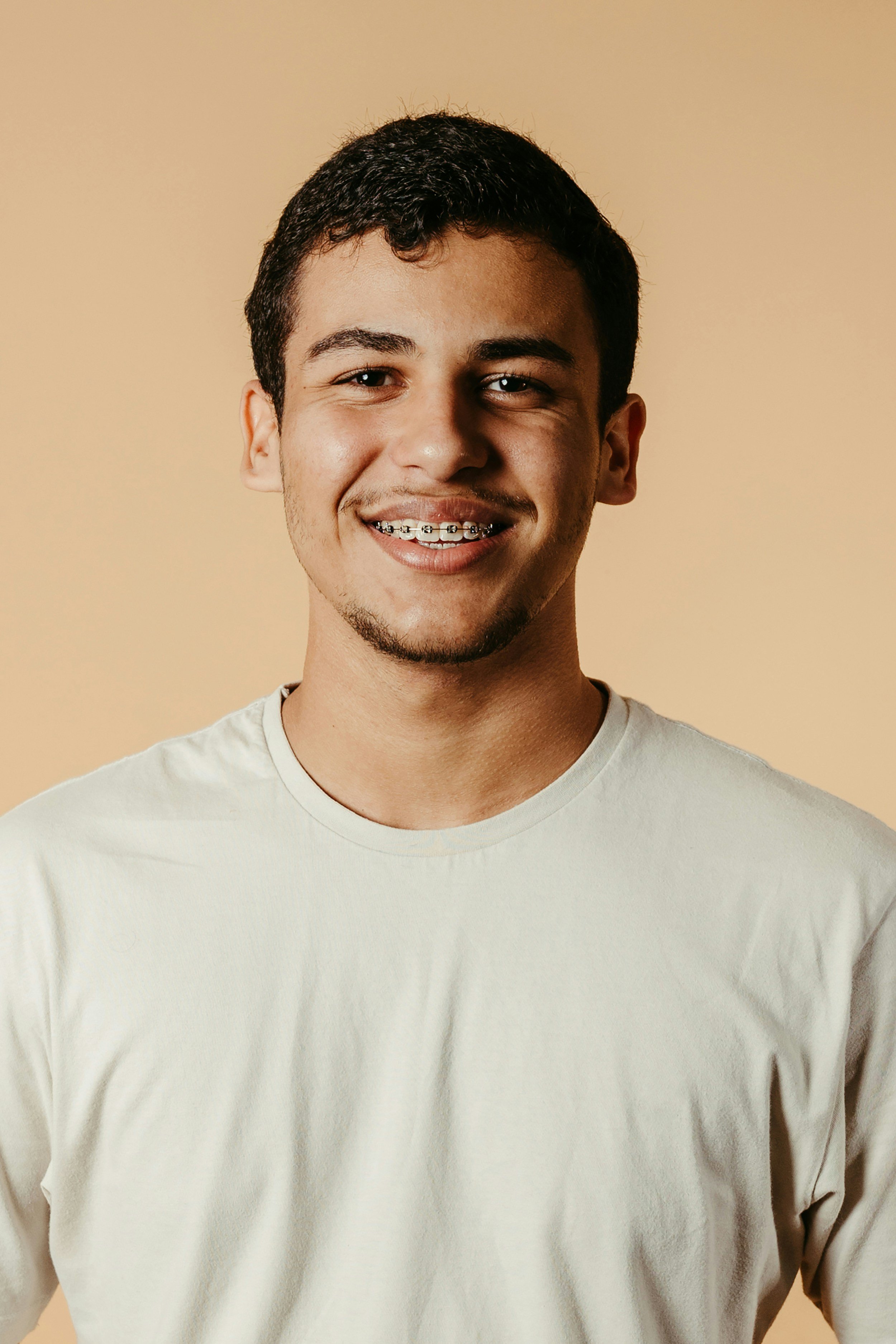 A young man smiling, wearing braces, with short dark hair, a light skin tone, in a plain white t-shirt, against a beige background.