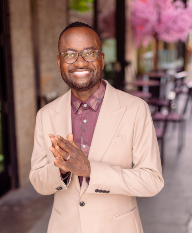 Smiling man with glasses in beige suit outdoors, pink blossoms in background.