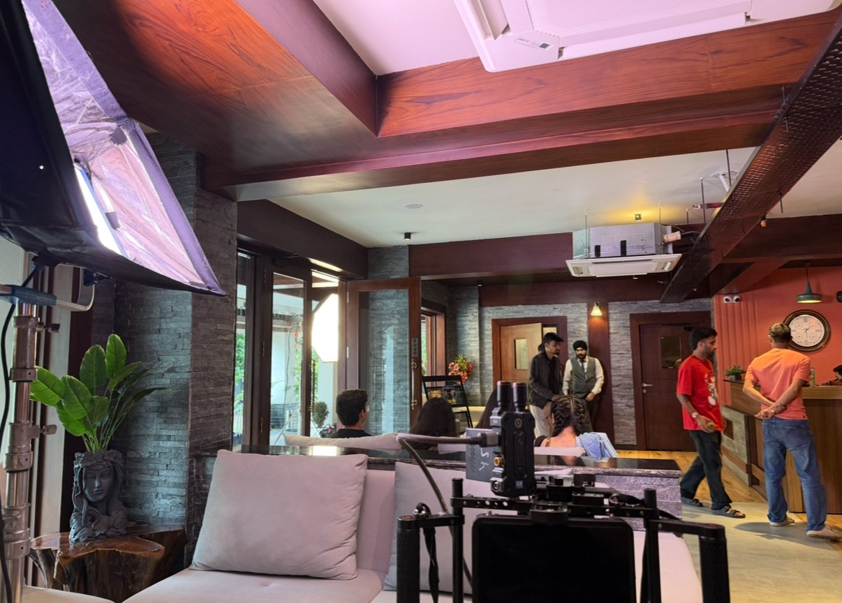 A lively indoor scene at a café or restaurant with people seated and standing, a large flat-screen TV on the left, modern decor including potted plants, wooden accents, brick walls, and a clock on the wall.