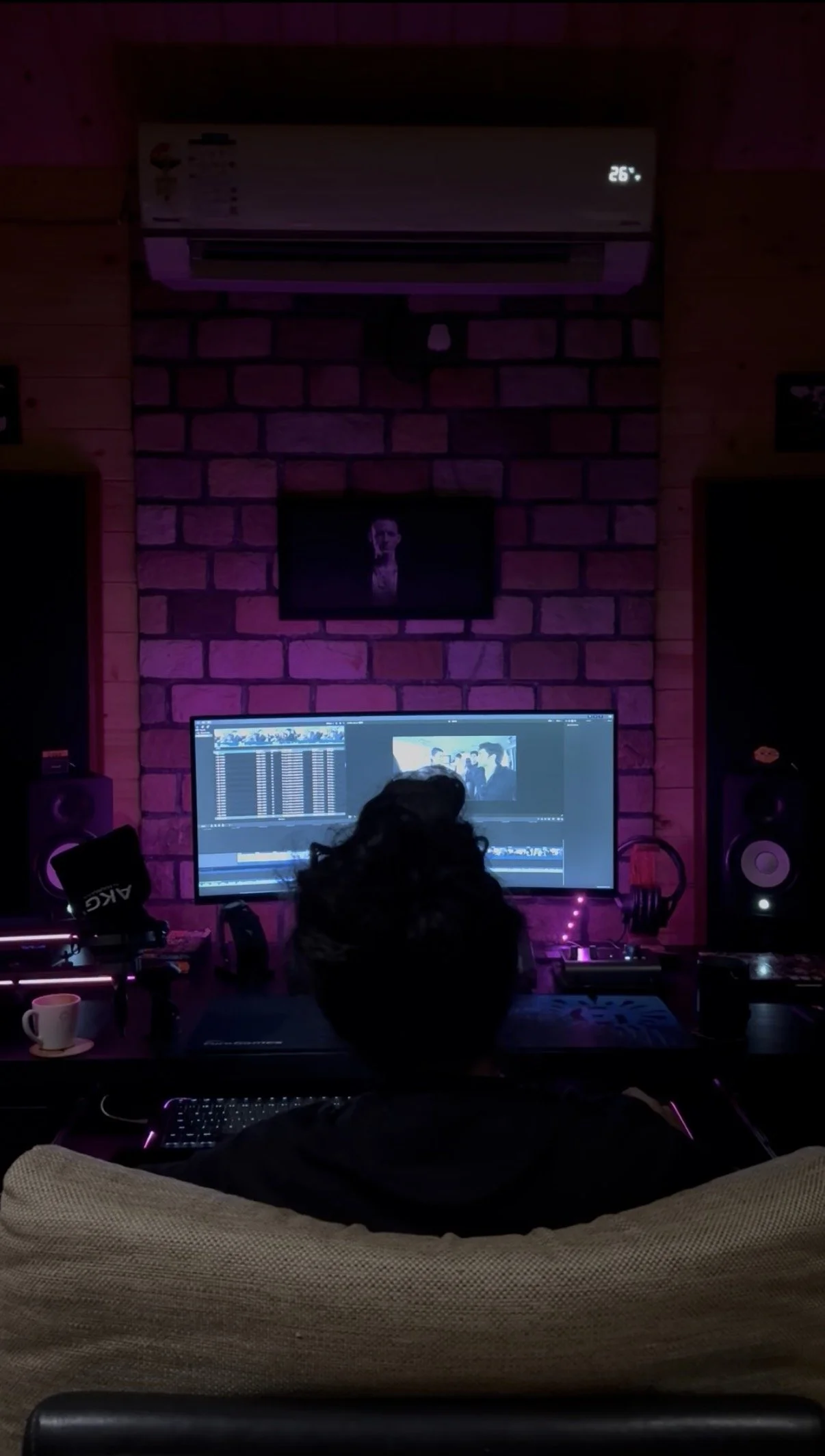 A person sitting on a couch in a music production studio, working on a computer with a large monitor displaying video editing software. Studio monitors and audio equipment are visible around the workspace, with purple ambient lighting and a brick wal