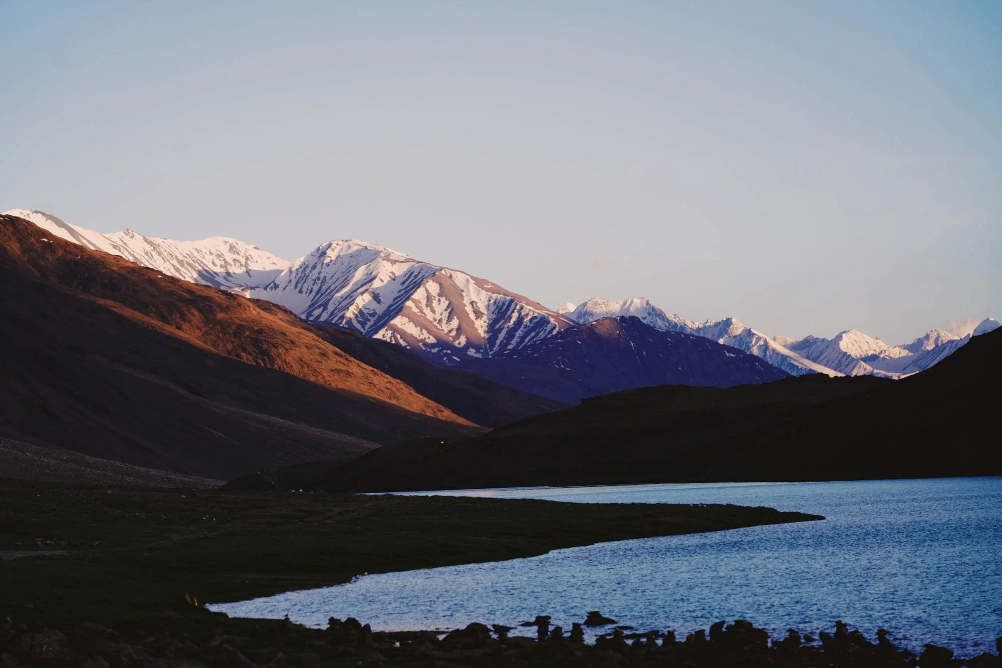 Mountain range with snow-capped peaks, a lake in the foreground, and a clear sky above.