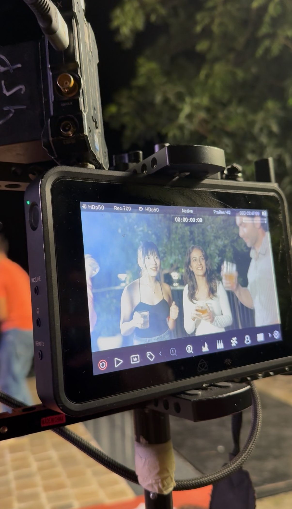 On a monitor screen, there are two women holding drinks at an outdoor event at night. A third person is partially visible on the right holding a drink.