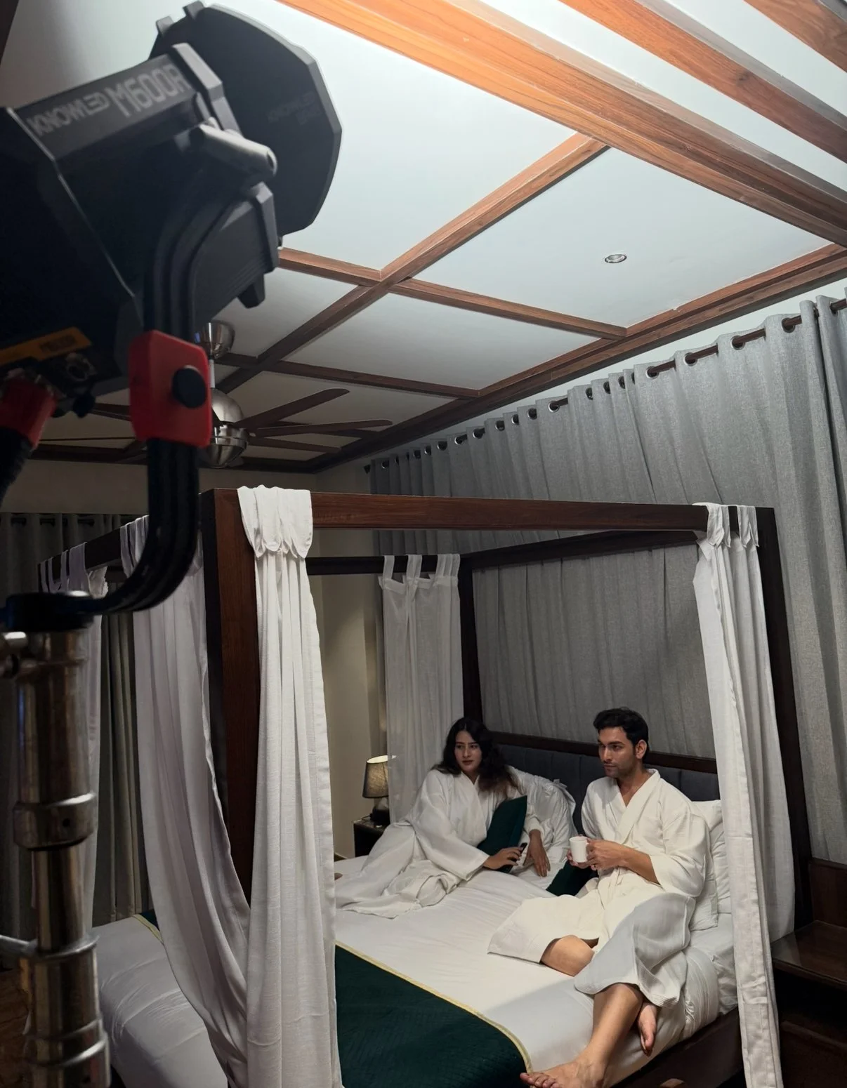 Two people in white robes sitting on a bed with a canopy, in a room with gray curtains and wooden ceiling panels, during a film shoot.