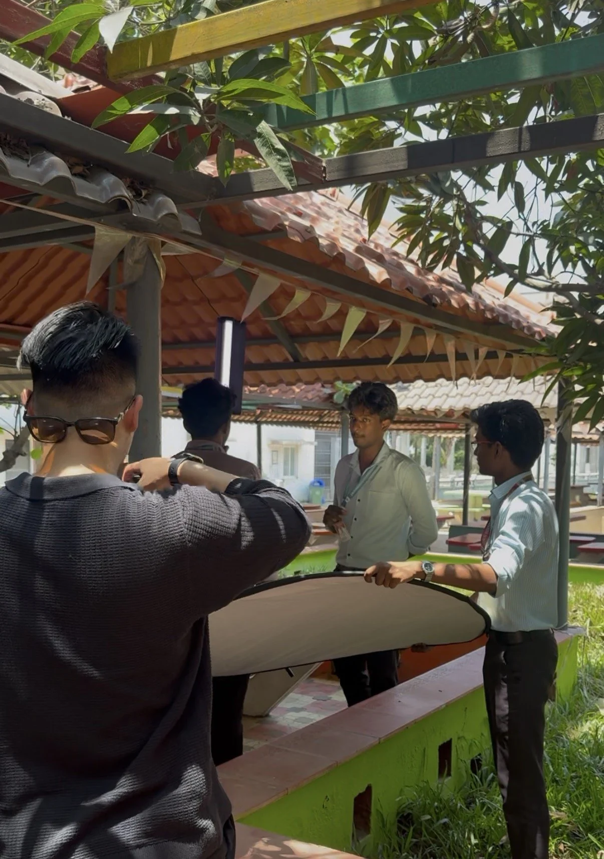 Four young men are outdoors under a shaded area with a tiled roof, surrounded by greenery and trees. Two of them are in conversation, one is holding a reflector, and the fourth is adjusting a camera. The setting appears to be a casual photo or video 