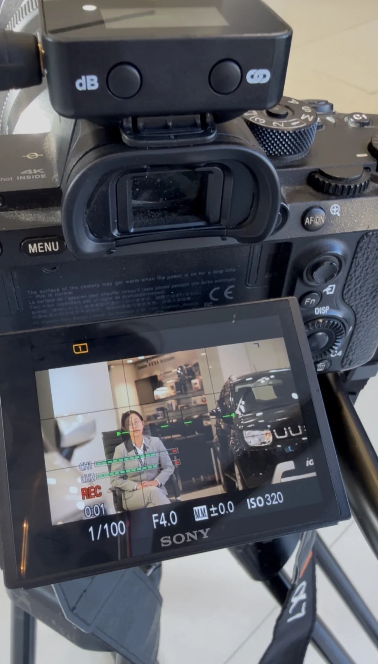 The image shows a DSLR camera with a monitor displaying a woman sitting in front of a camera in a studio, with some indoor cars in the background.