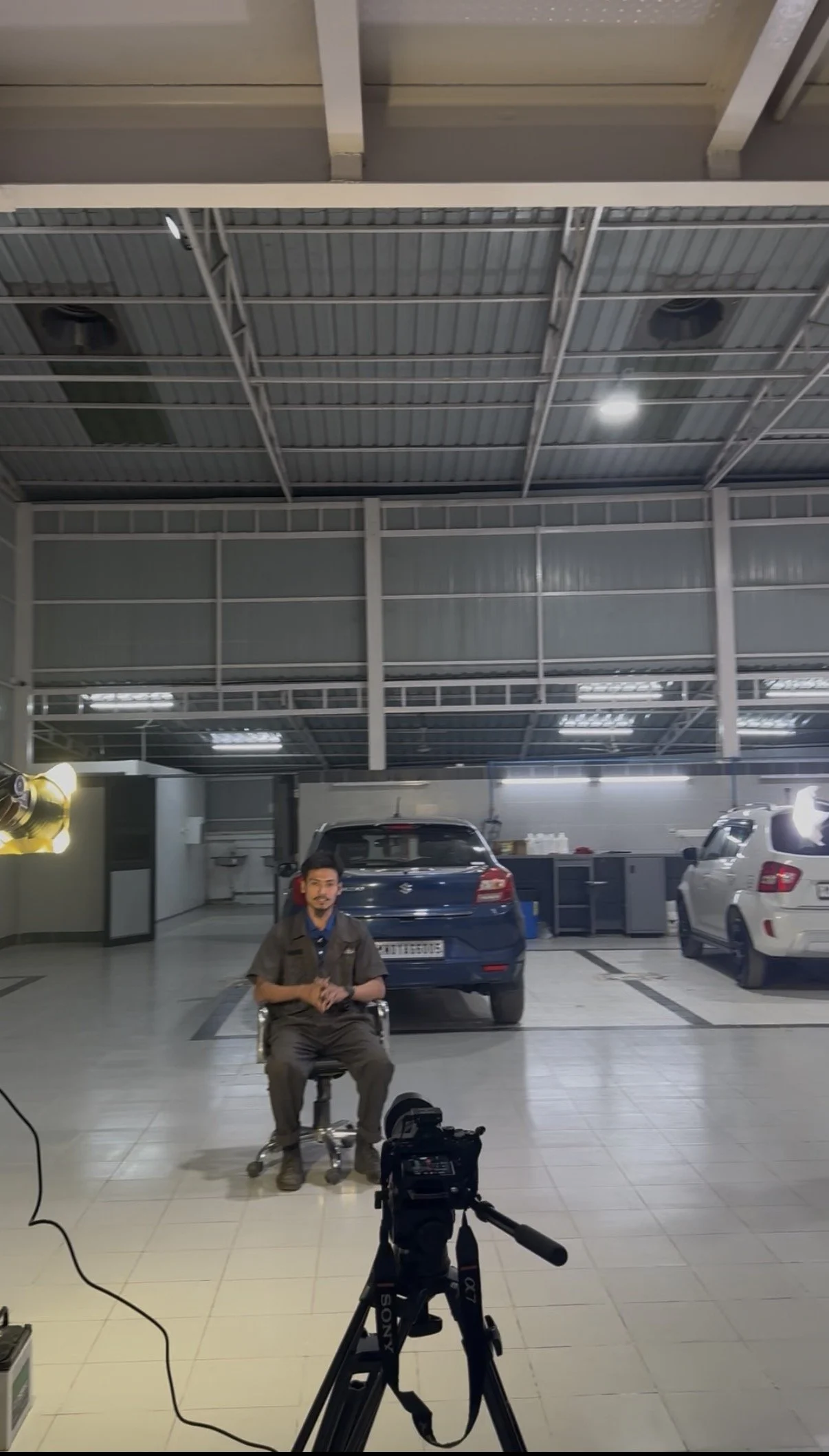 An indoor parking garage with a man sitting on a chair, a camera on a tripod in front of him, and four parked cars behind him. The ceiling is made of metal with industrial lighting.