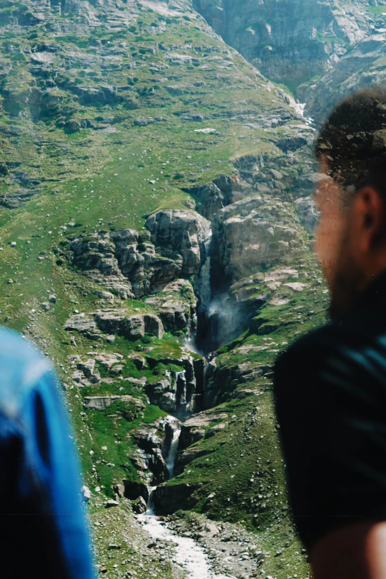 People observing a mountain landscape with a waterfall, rocky terrain, and green hills, through a glass window.