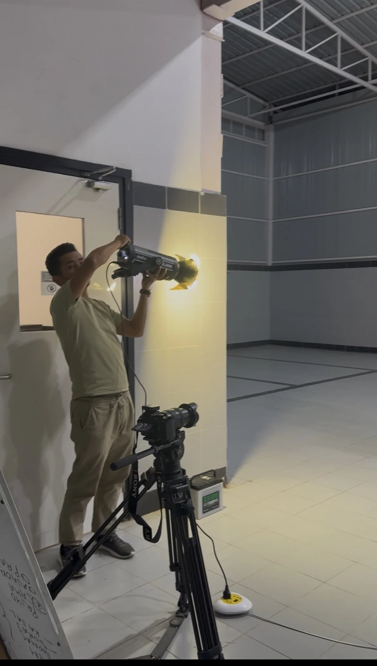 A person operating a handheld device, possibly a laser scanner, in an indoor industrial or warehouse setting. There is a camera on a tripod and other equipment nearby, and the scanner emits a yellow light. The background shows metal beams and a wall.