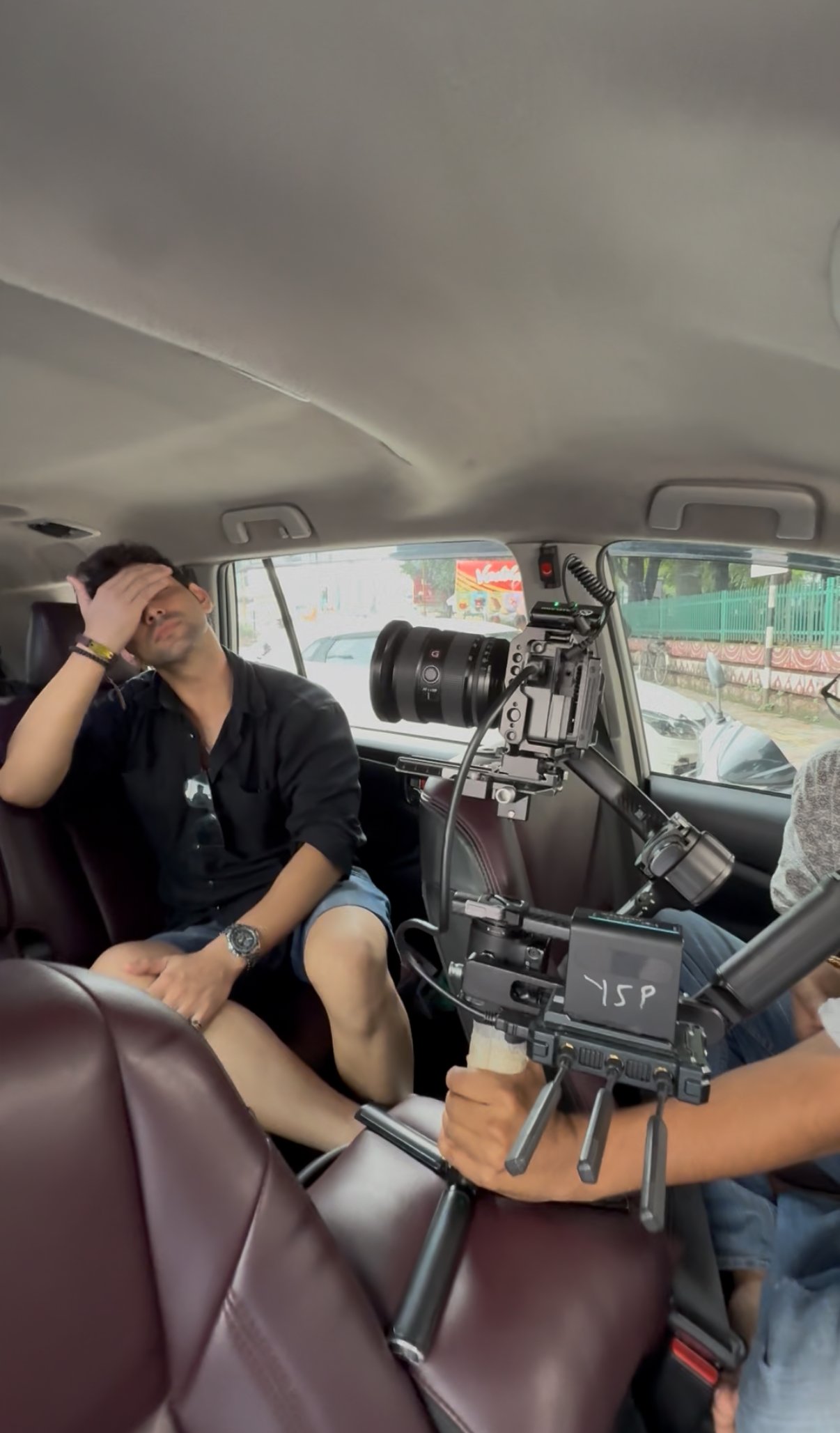 A man sitting in the back of a vehicle with a film camera mounted on a stabilizer, while another man appears to be behind the camera, filming him. The man in the back has his hand on his forehead, and there is a city scene visible through the vehicle