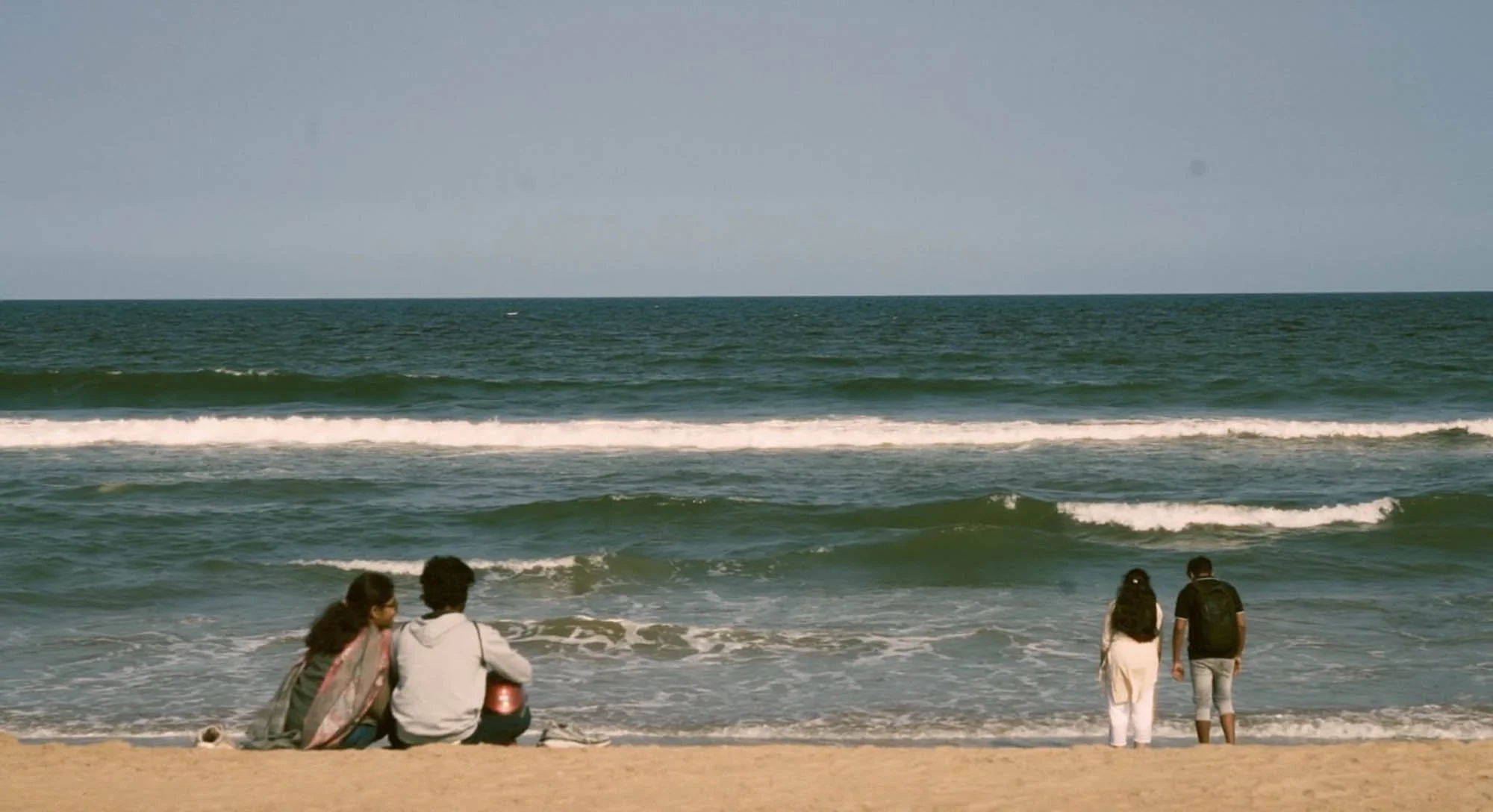 People sitting and standing on a sandy beach near the water, with ocean waves and a cloudy sky in the background.