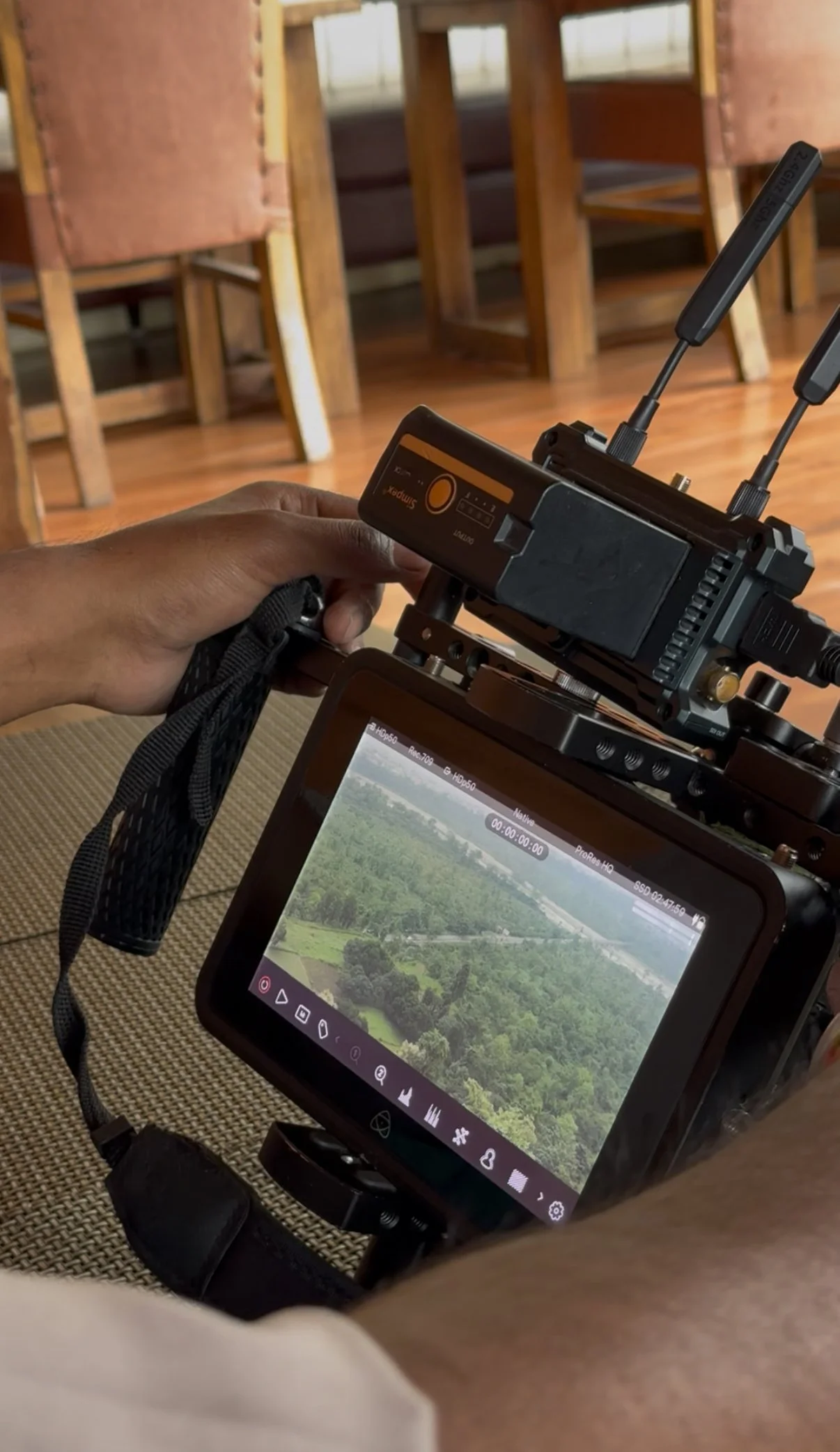 A person holds a camera monitor displaying an aerial view of trees and greenery inside a room with wooden chairs.
