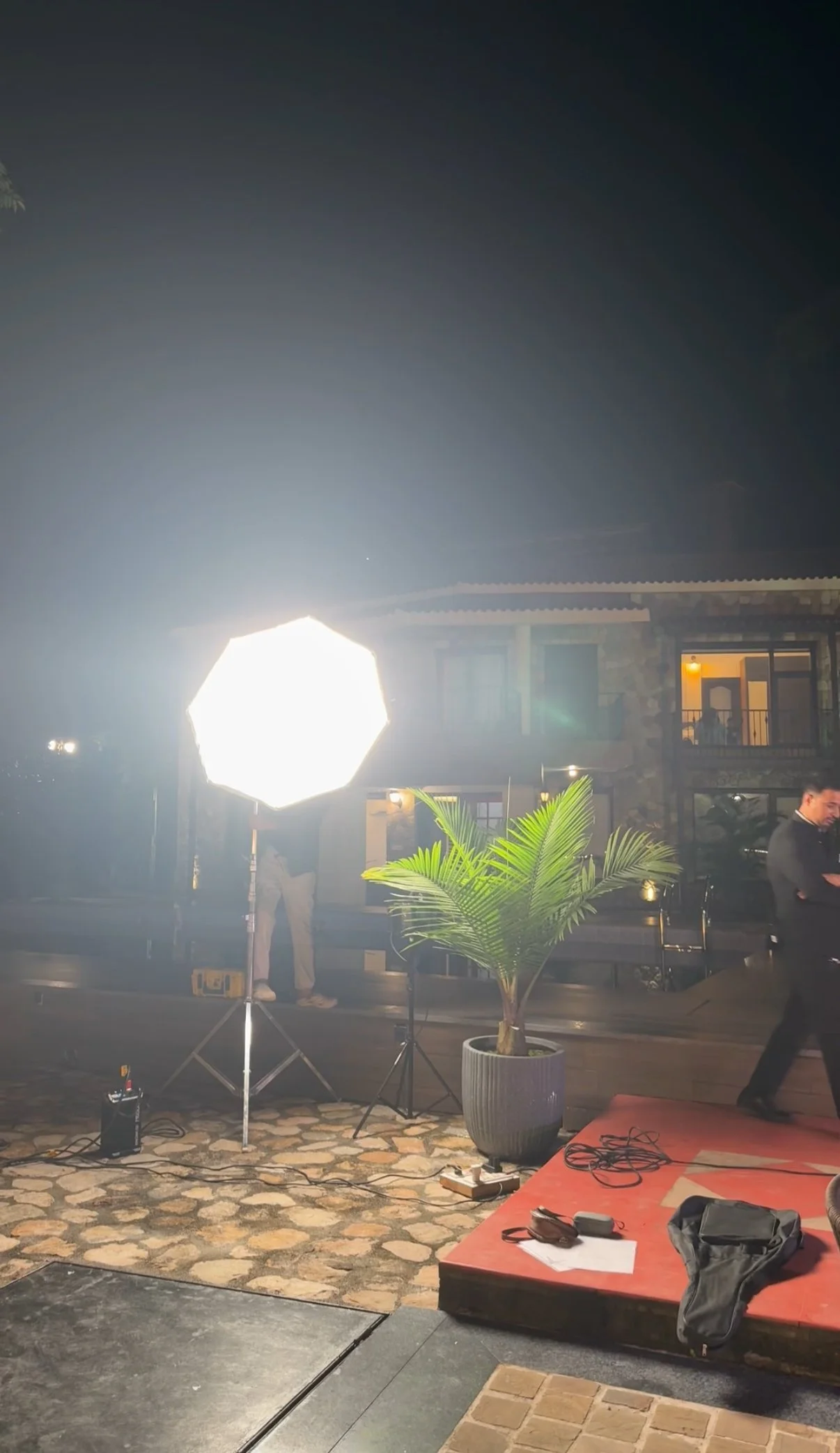 Nighttime outdoor filming setup with professional lighting, a potted plant, and people in the background near a building with balconies.