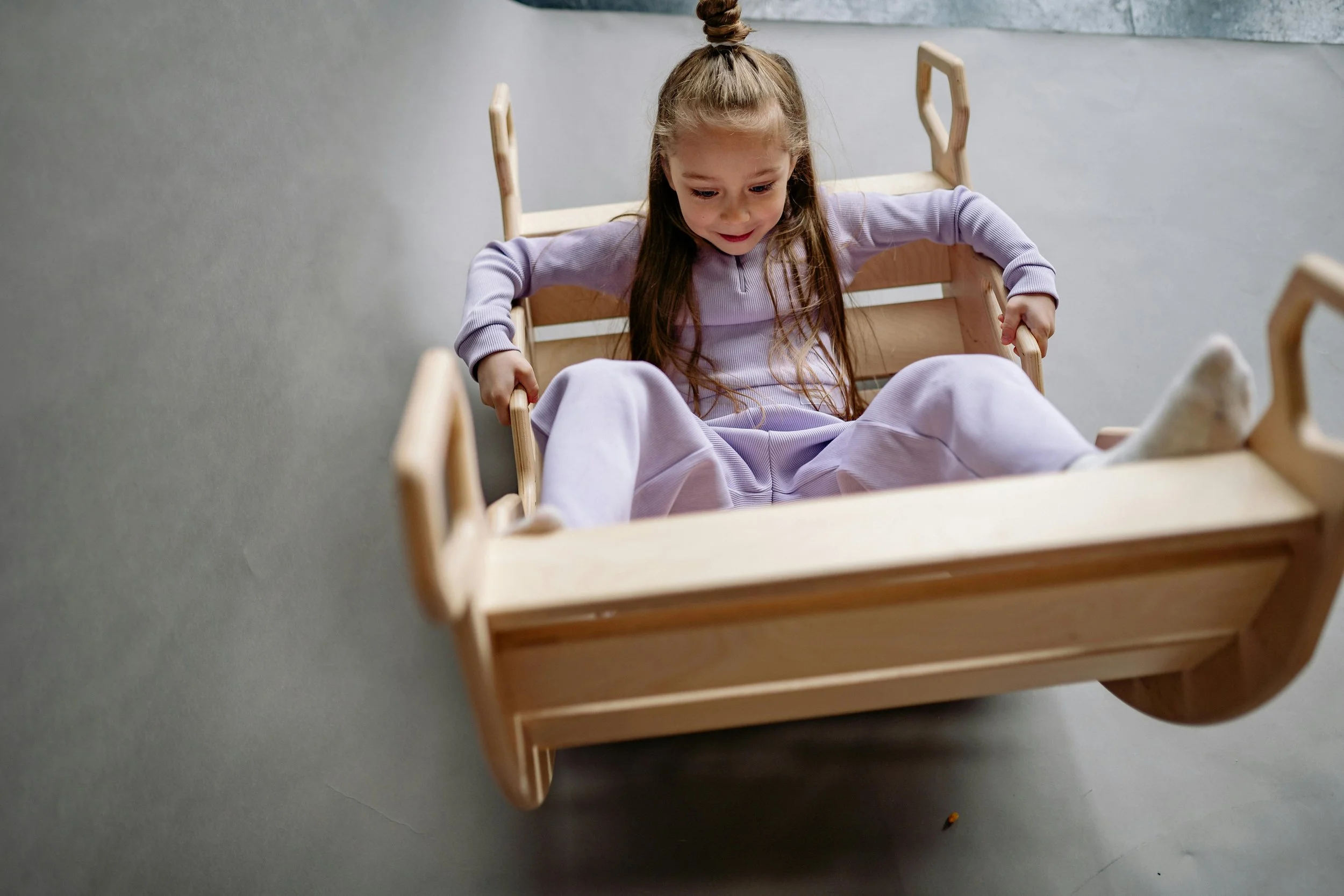 A young girl wearing a lavender outfit sitting in a wooden wagon, holding its sides with a playful expression.