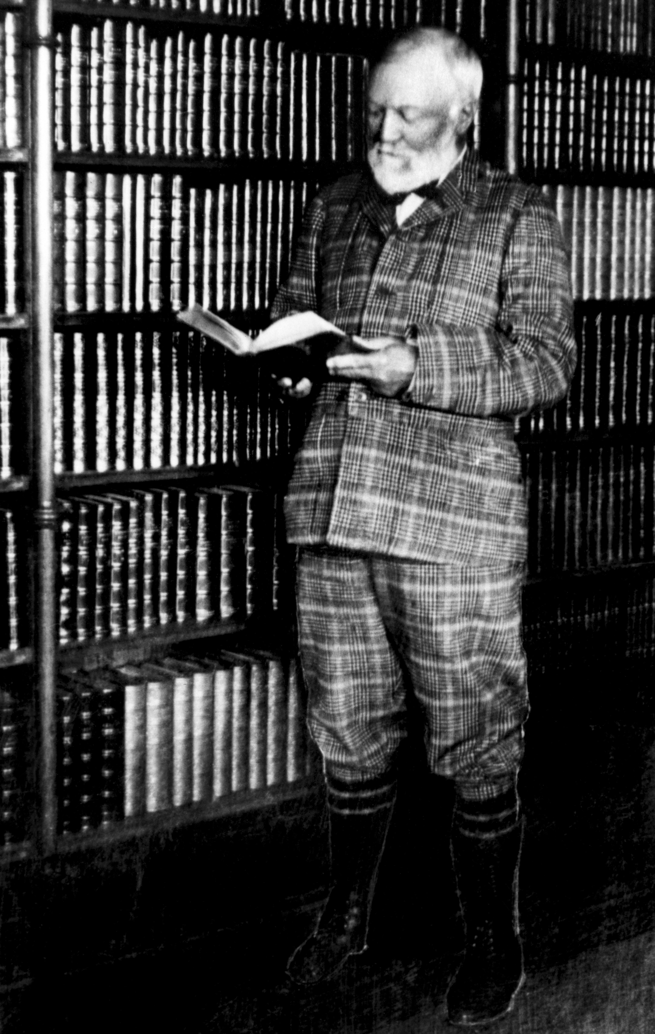 An elderly man with a beard and glasses is reading a book in a library filled with bookshelves.