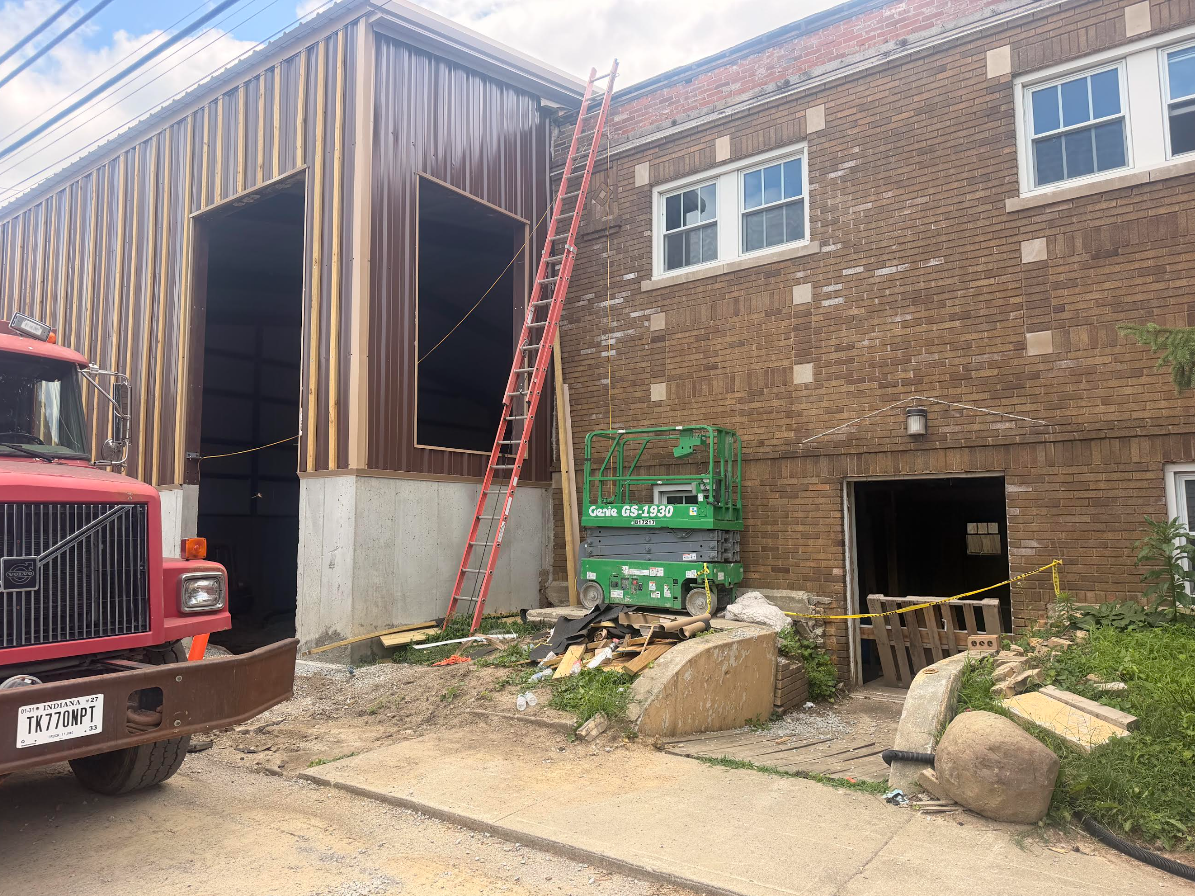 Construction site with a two-story brick building and a partially built addition with metal siding, a red ladder leaning against the building, a green lift, construction materials, debris and a red vehicle in the foreground.