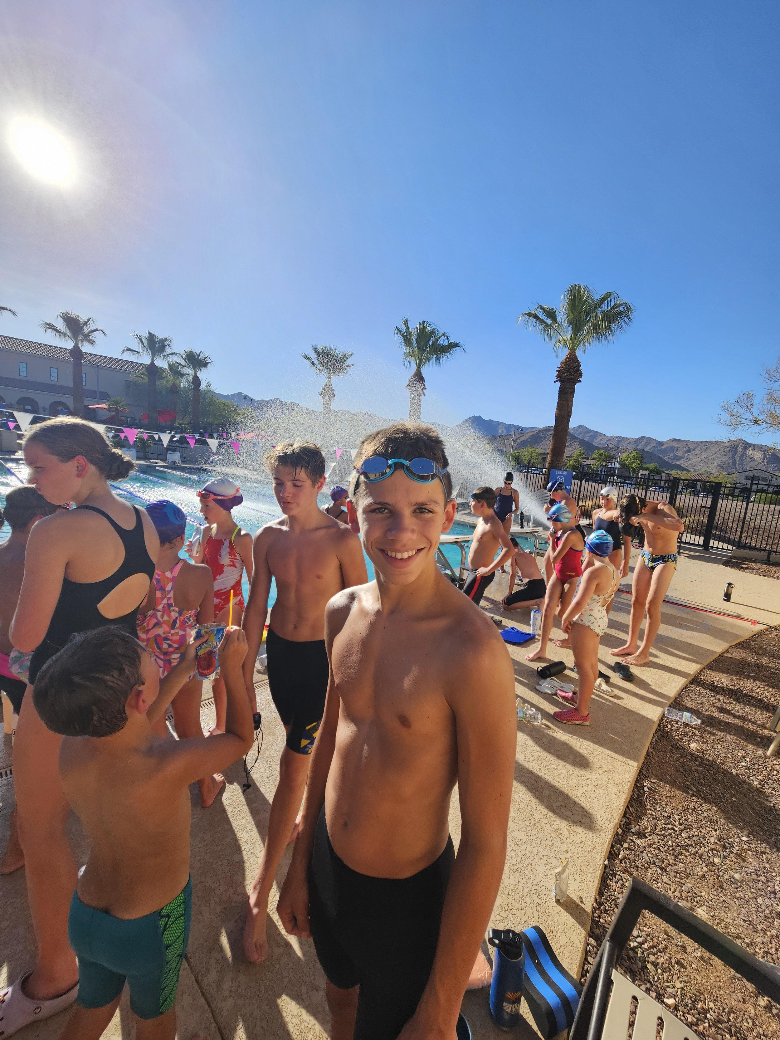 Children at an outdoor swimming pool, some with swim caps and goggles, in a sunny desert landscape with mountains and palm trees in the background.