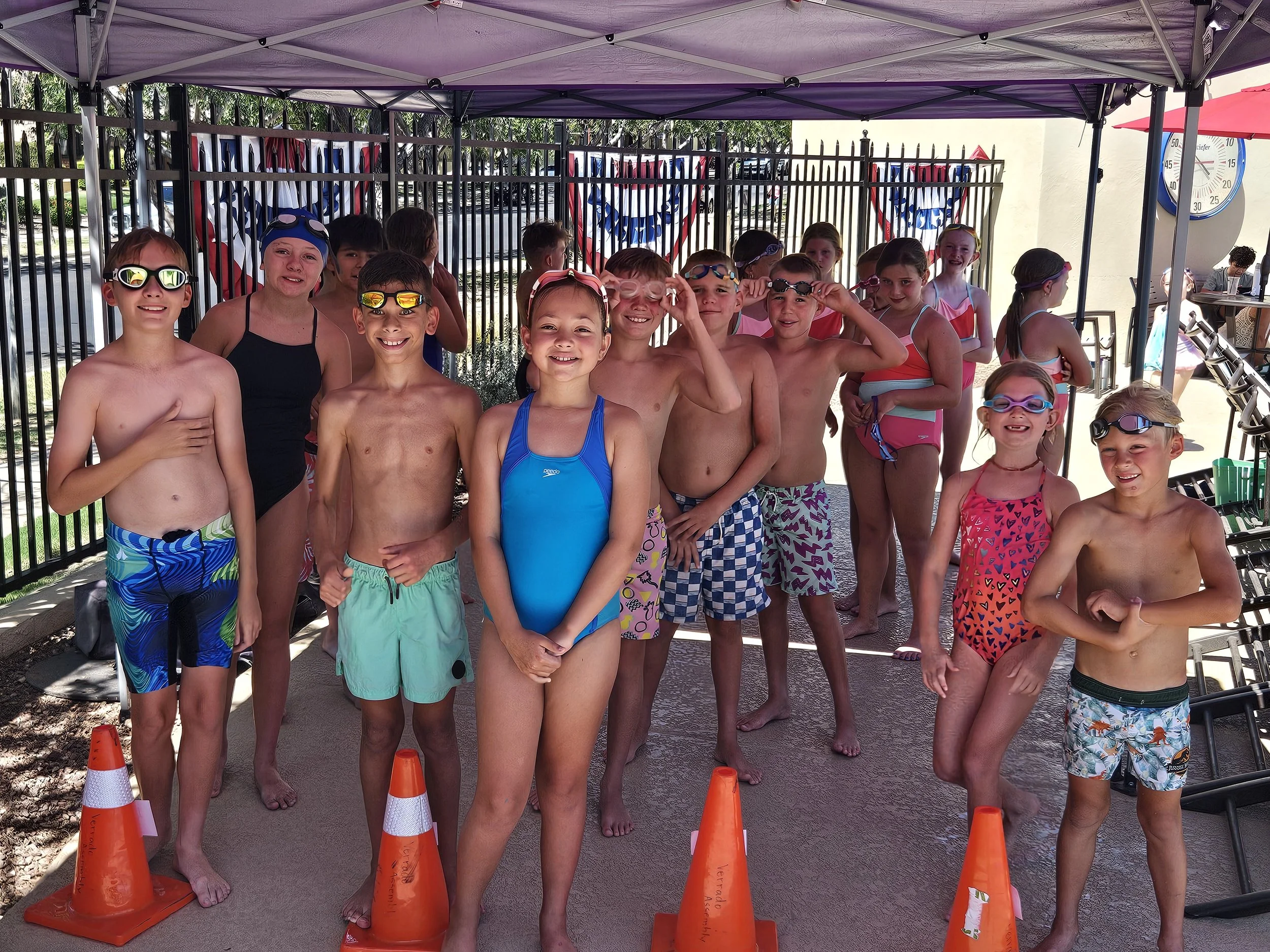 A group of children in swimsuits and goggles standing behind orange traffic cones under a canopy outdoors, with some children smiling and posing for the photo.