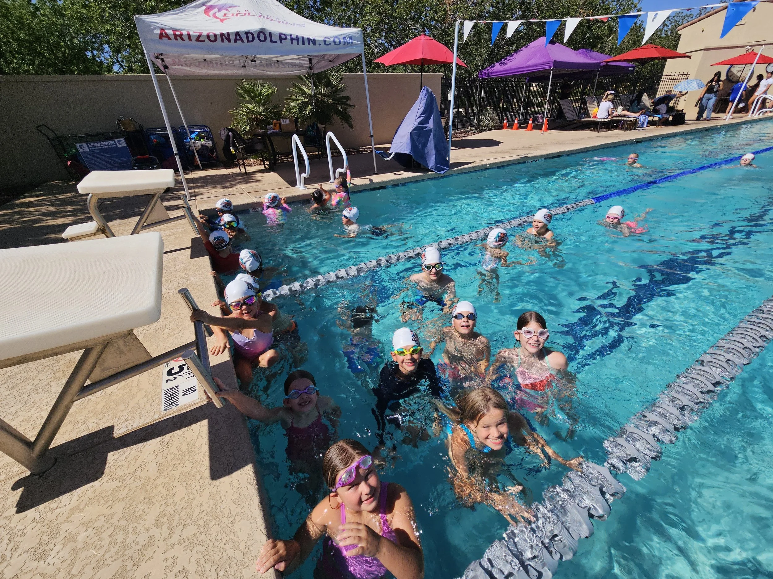 Children wearing swim caps and goggles enjoying a swim lesson in an outdoor pool under a sunny sky, with umbrellas and chairs on the poolside.