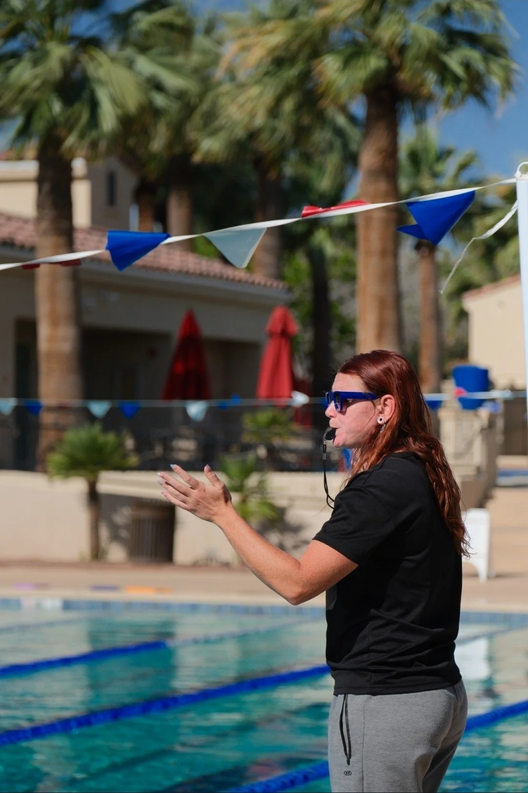 A swimming coach wearing sunglasses and a whistle giving instructions by a swimming pool with colorful bunting and palm trees in the background.