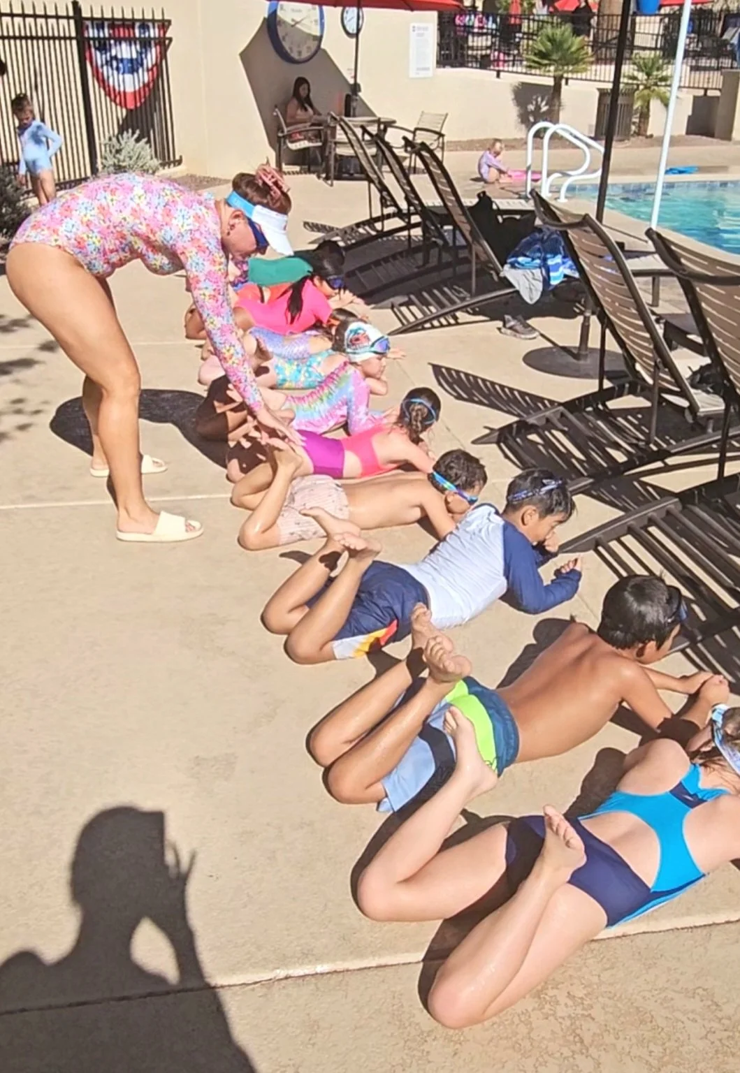 Children lying on the ground at a poolside, in a line holding each other's feet, with an adult supervising nearby.