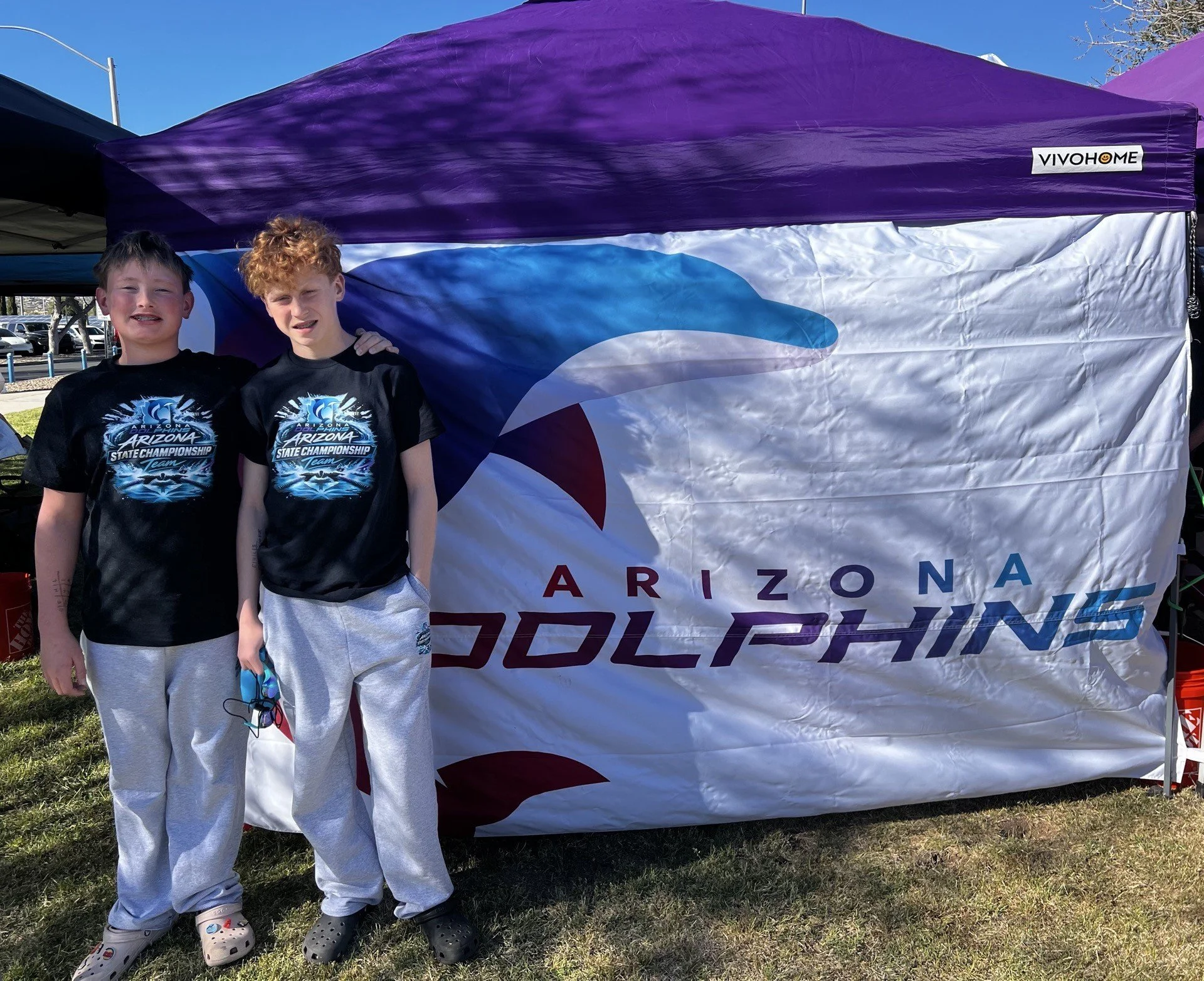 Two boys wearing black t-shirts with blue graphics and gray sweatpants, standing outdoors in front of a large flag and purple canopy at a sports event. The flag displays the word "Arizona" and a stylized eagle or bird logo.