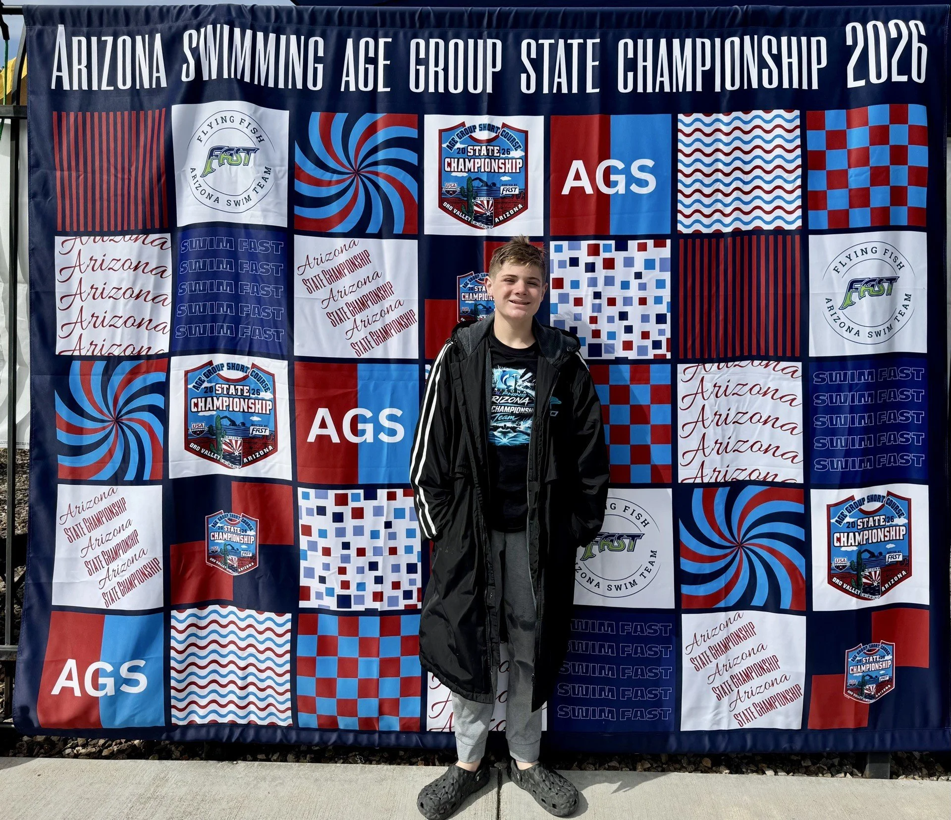 A young boy standing in front of a large banner displaying various patriotic and sports logos, text, and patterns related to the Arizona Swimming Age Group State Championship 2026. The boy is smiling, wearing a black jacket over a blue t-shirt, gray 