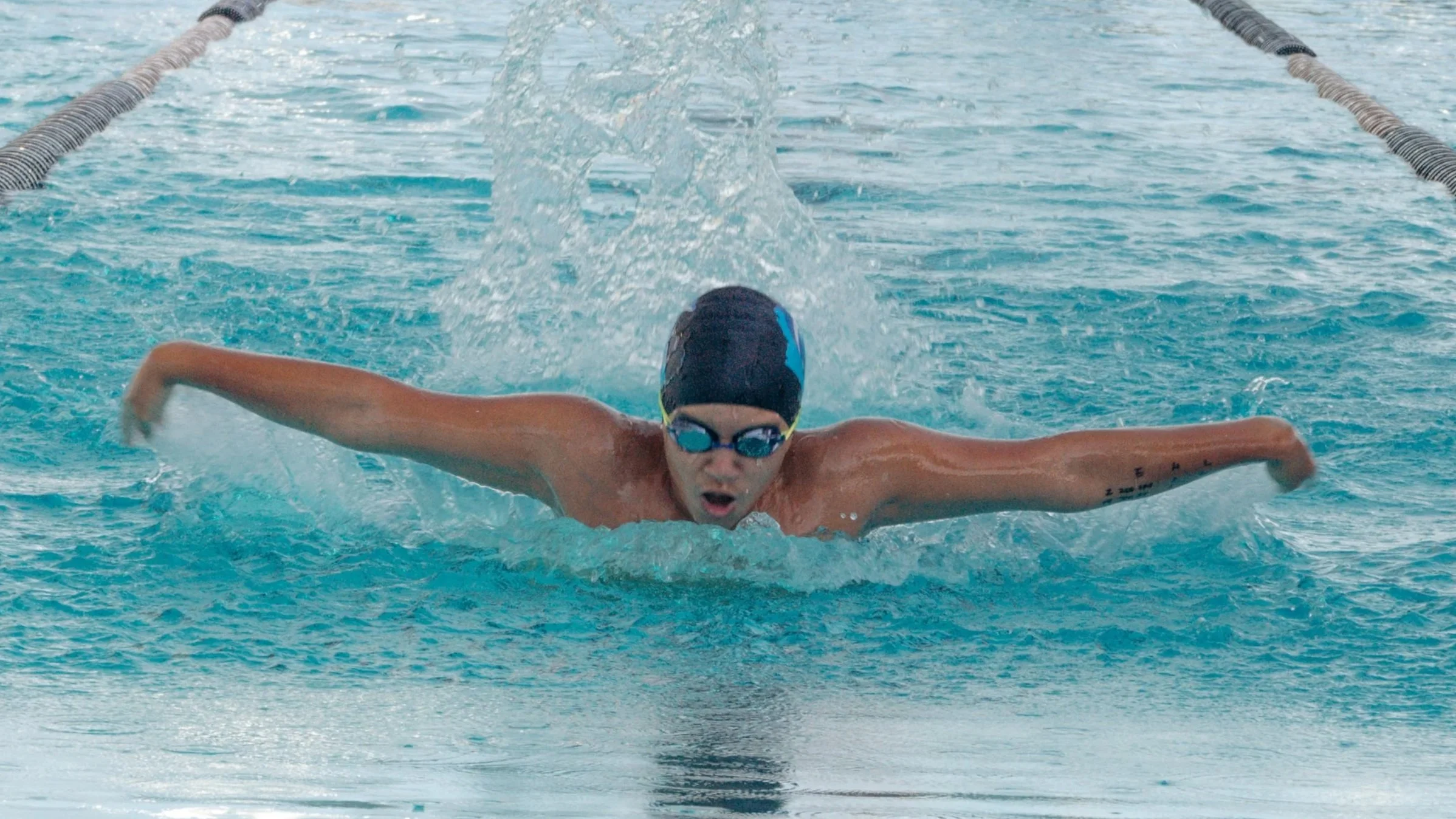 Swimmer performing a butterfly stroke in a swimming pool, wearing goggles and a swim cap.