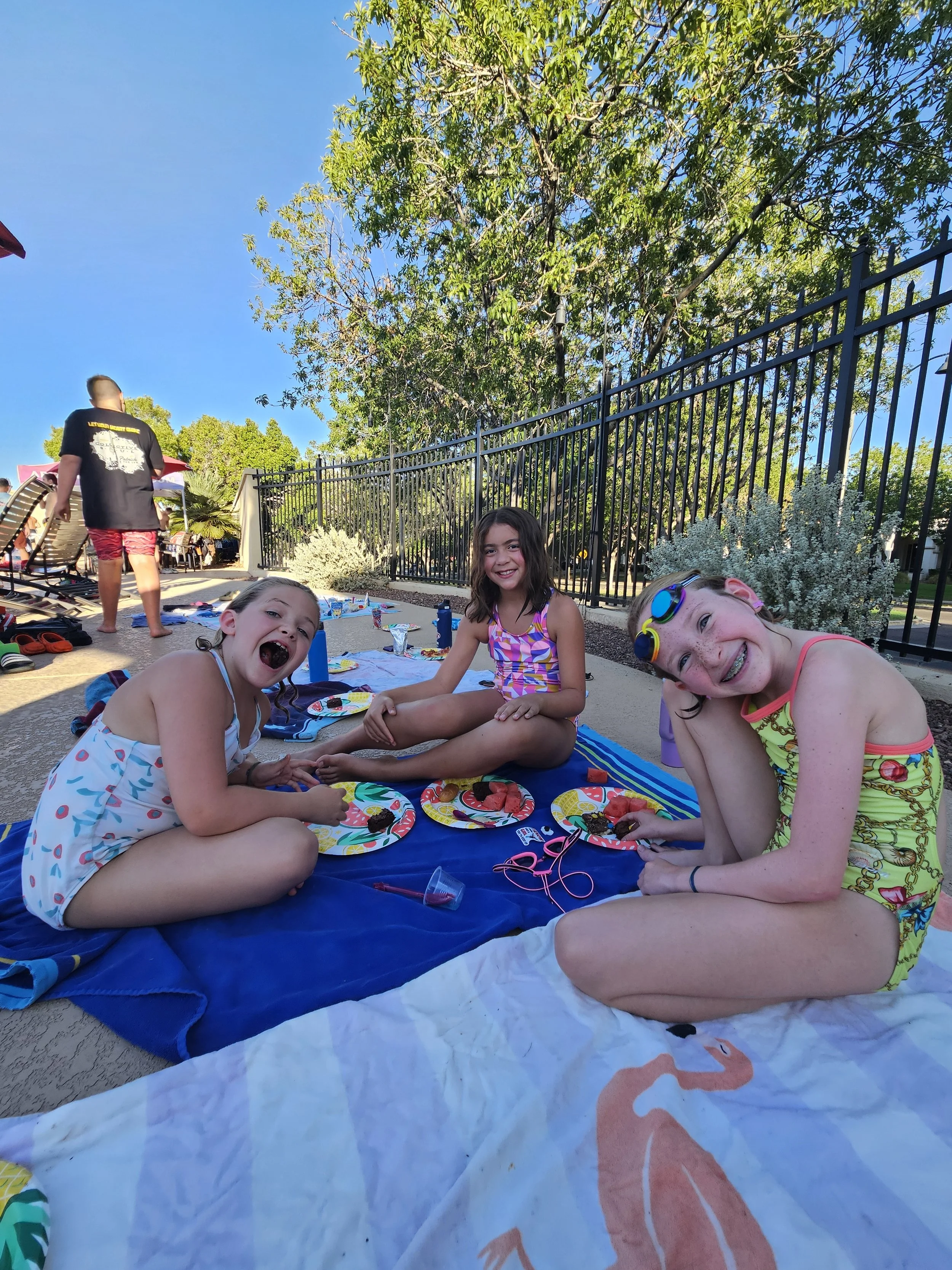 Three young girls are sitting on a blanket at an outdoor poolside area, smiling and enjoying snacks. Two boys are in the background, with one walking away and the other partially visible. There is a black fence, trees, and a clear blue sky.