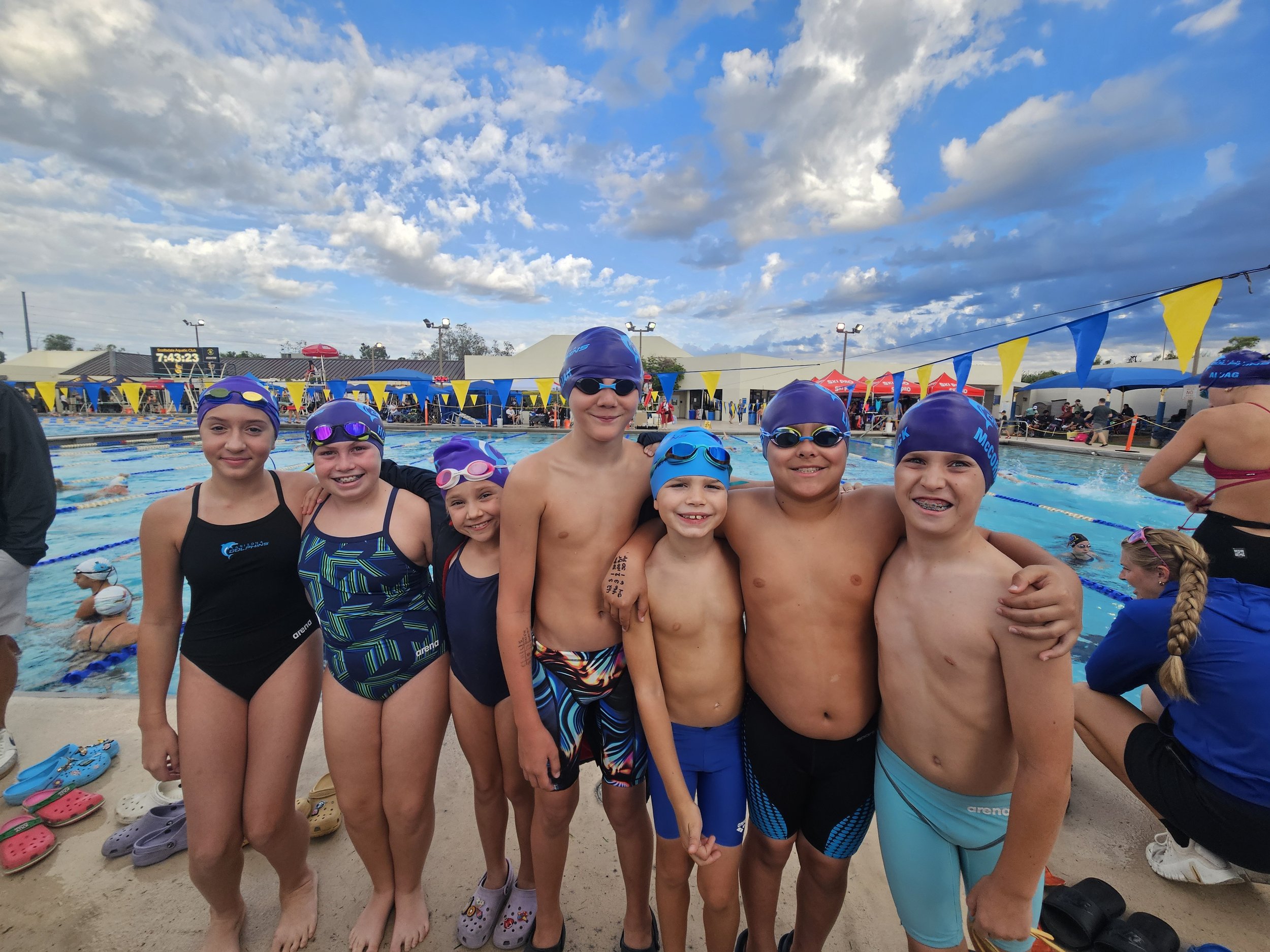 Group of young swimmers, both boys and girls, standing together at an outdoor swimming pool, smiling and wearing swim caps and goggles, with a pool and cloudy sky in the background.