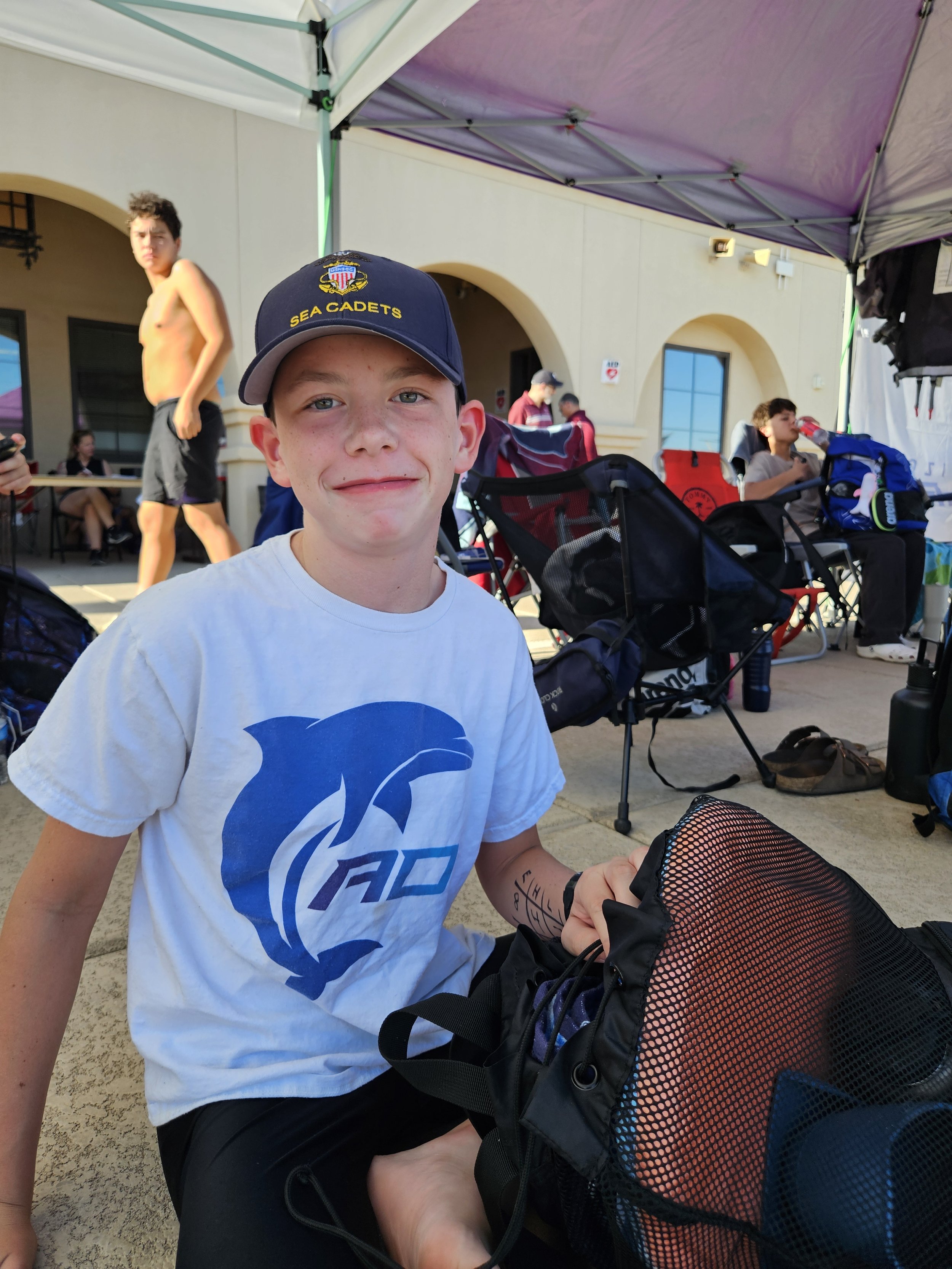 A young boy wearing a navy blue 'Sea Cadets' baseball cap and a white T-shirt with a blue logo, sitting outdoors under a purple canopy. In the background, other children and teenagers are visible, some sitting and others standing, some drinking water, with sports bags and chairs around.