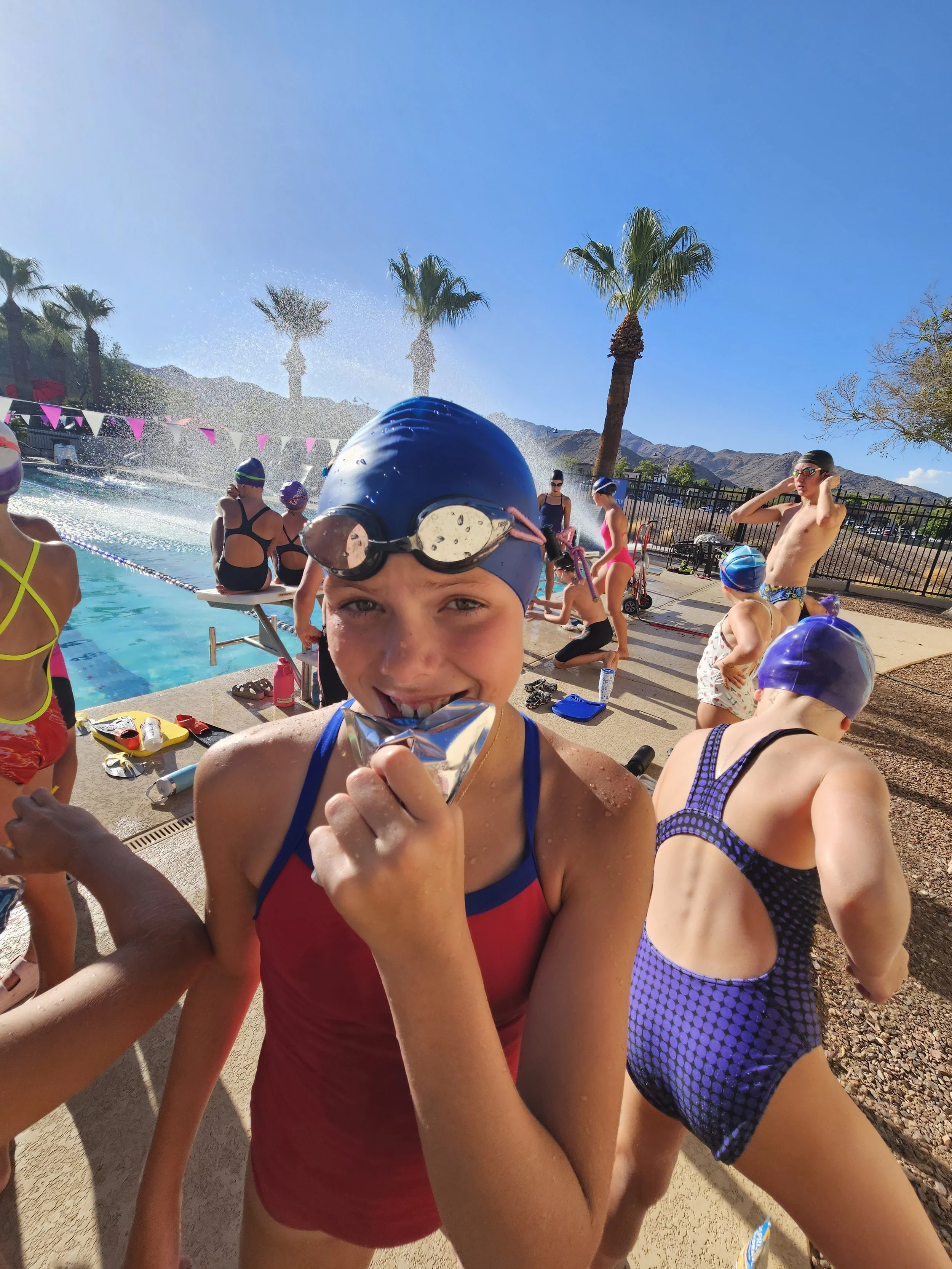 Children wearing swimsuits and swim caps at an outdoor pool, with palm trees and mountains in the background.